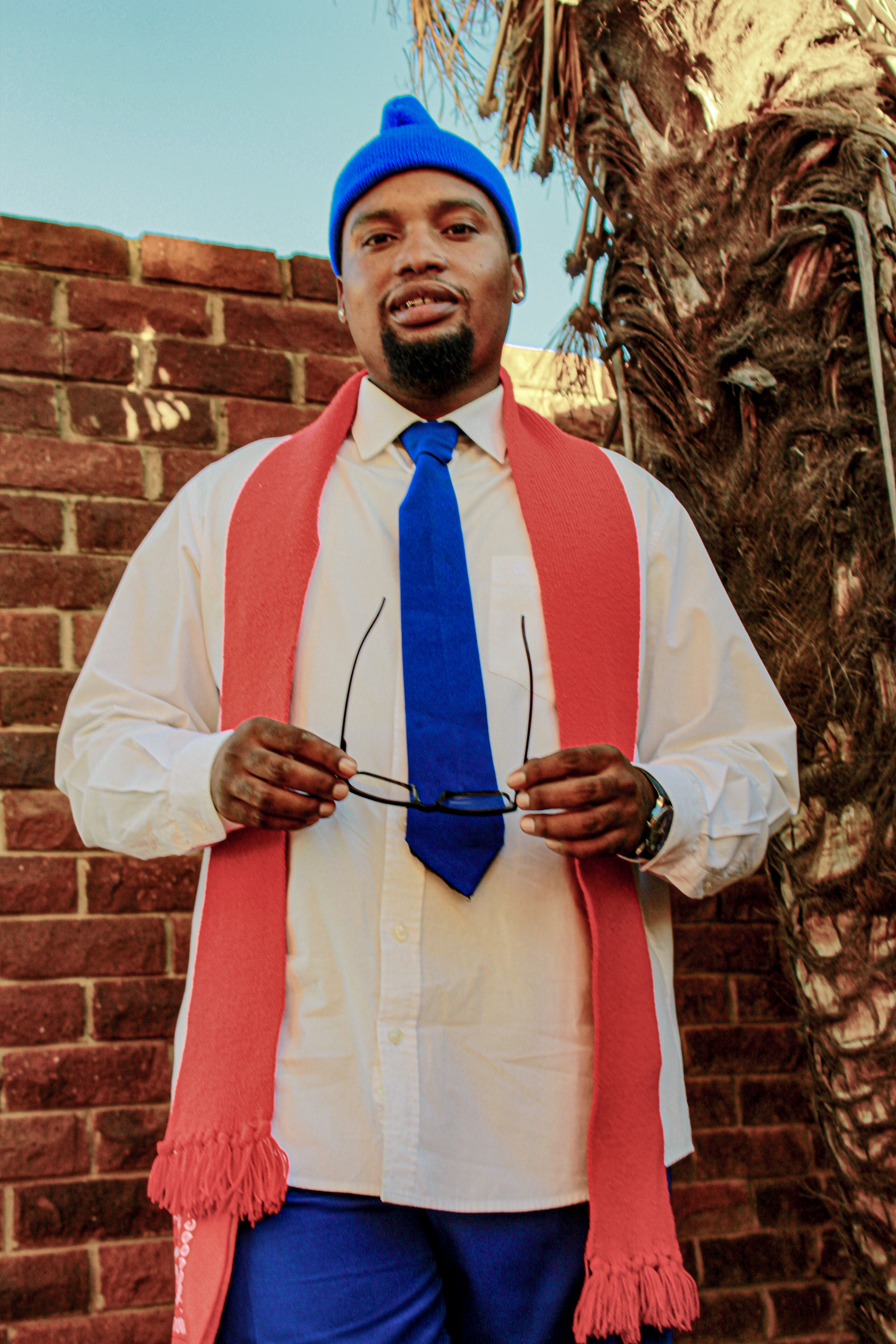 A man wearing a red scarf and blue hat stands confidently against a brick wall, exuding style.