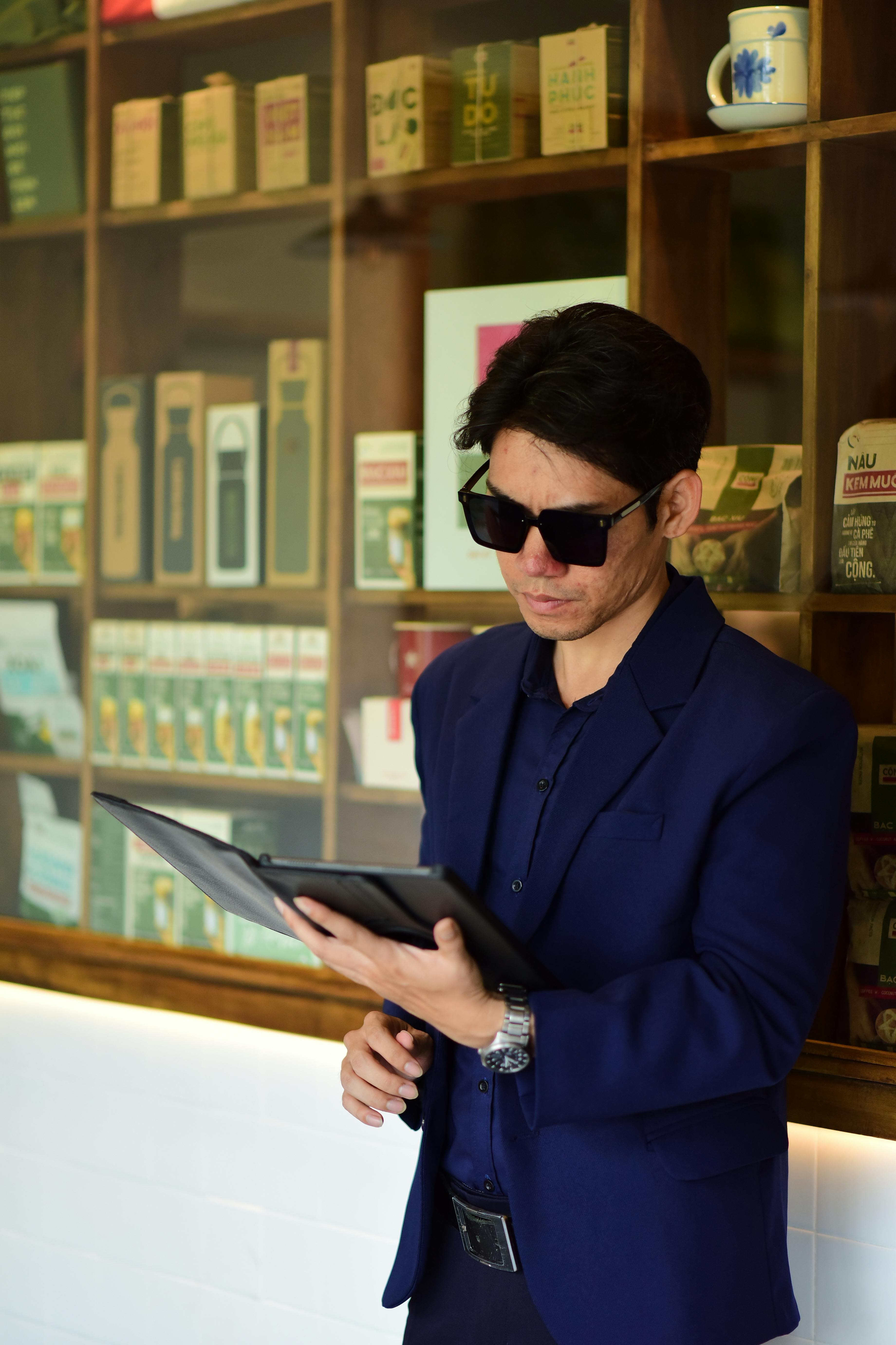 Gratis Un uomo elegante in abito blu legge su un tablet in un bar moderno con scaffali pieni di prodotti. Foto a disposizione