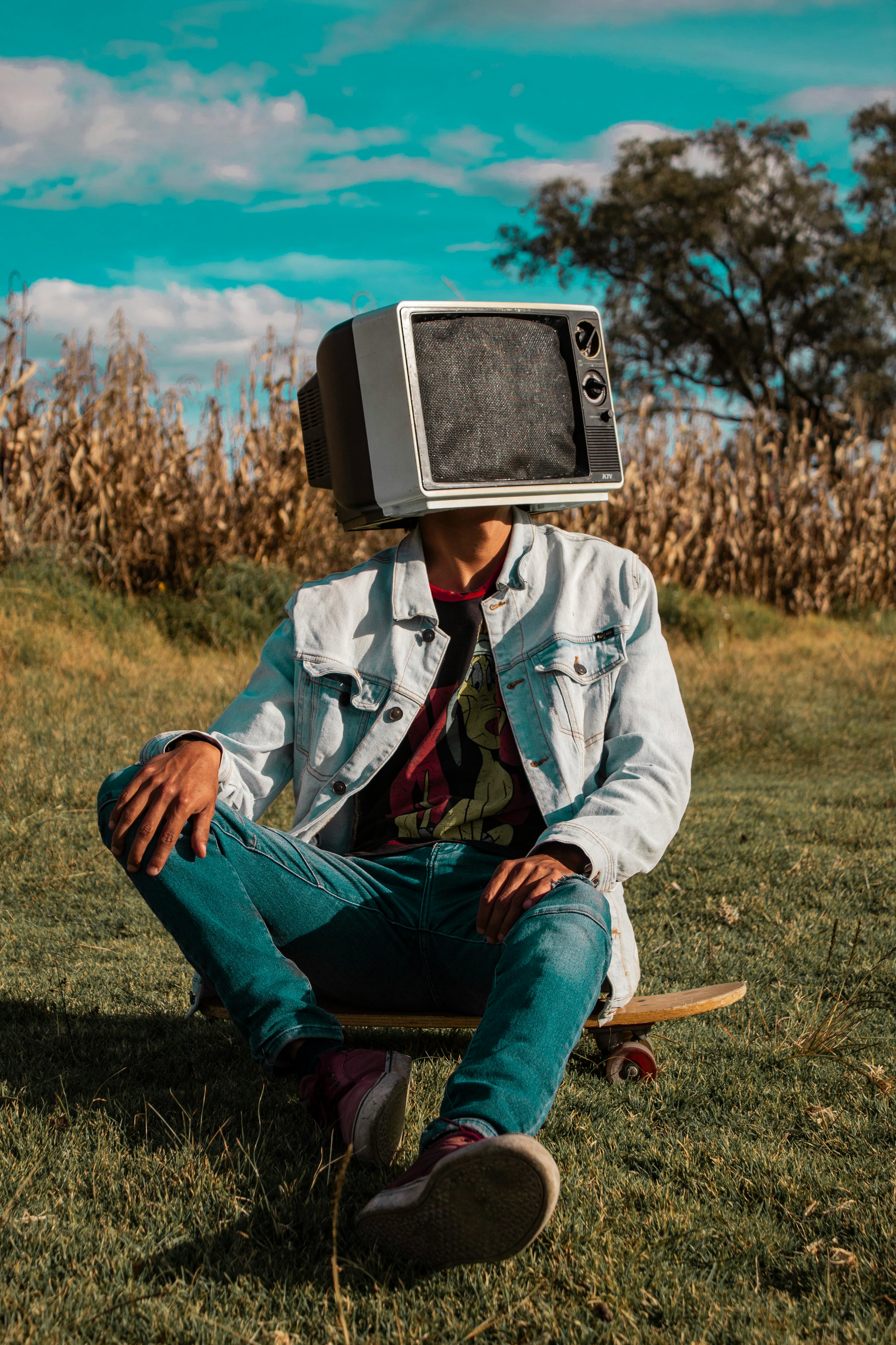 Gratis Seseorang dengan kepala berbentuk TV retro duduk di atas skateboard di sebuah lapangan. Foto Stok