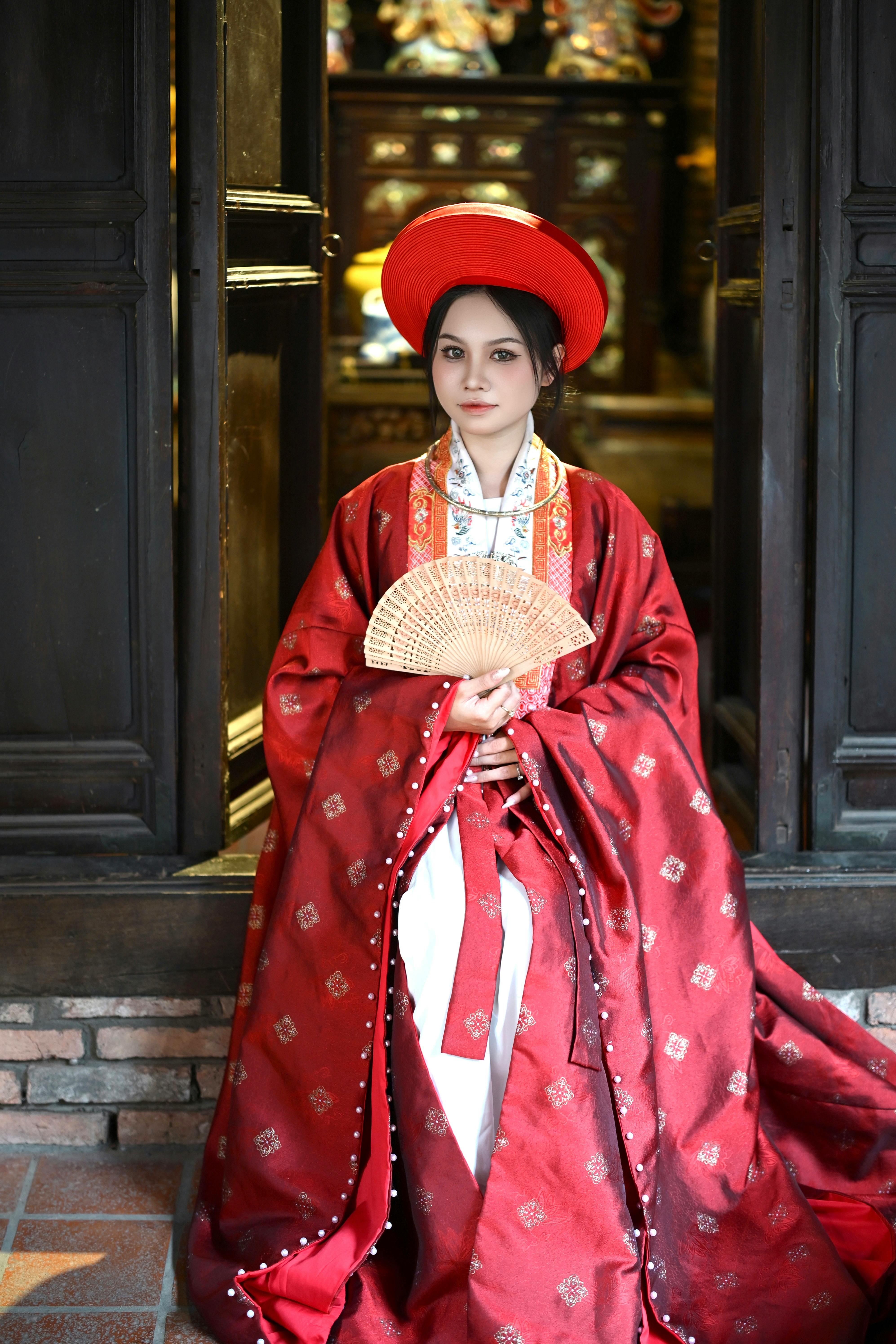 Young Asian woman in traditional red hanfu, holding fan indoors.