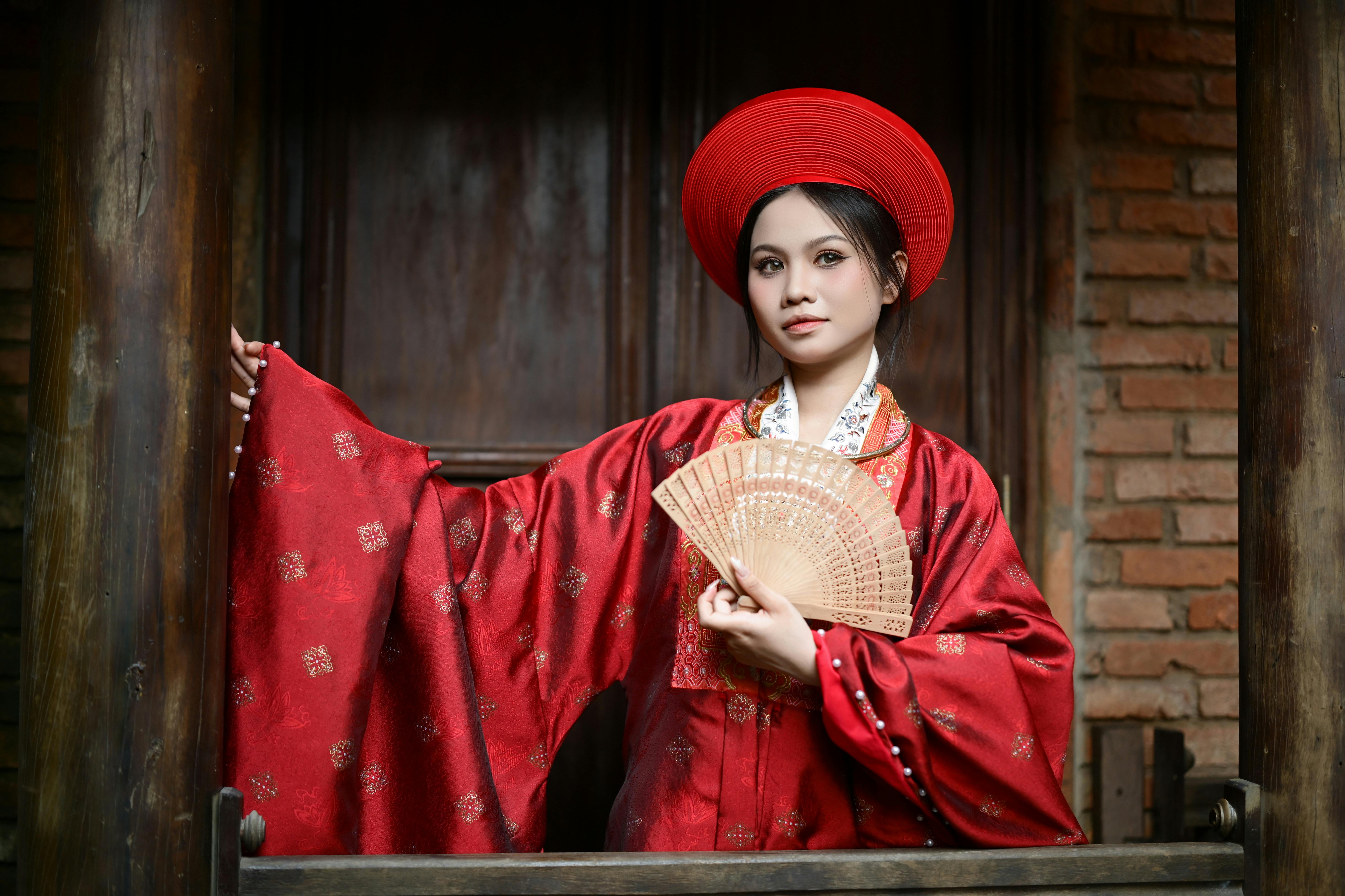 Gratis Una mujer joven con el atuendo rojo tradicional sosteniendo un abanico, de pie con gracia al aire libre. Foto de stock