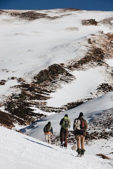Three hikers journey through snow-covered mountain terrain under a clear blue sky.