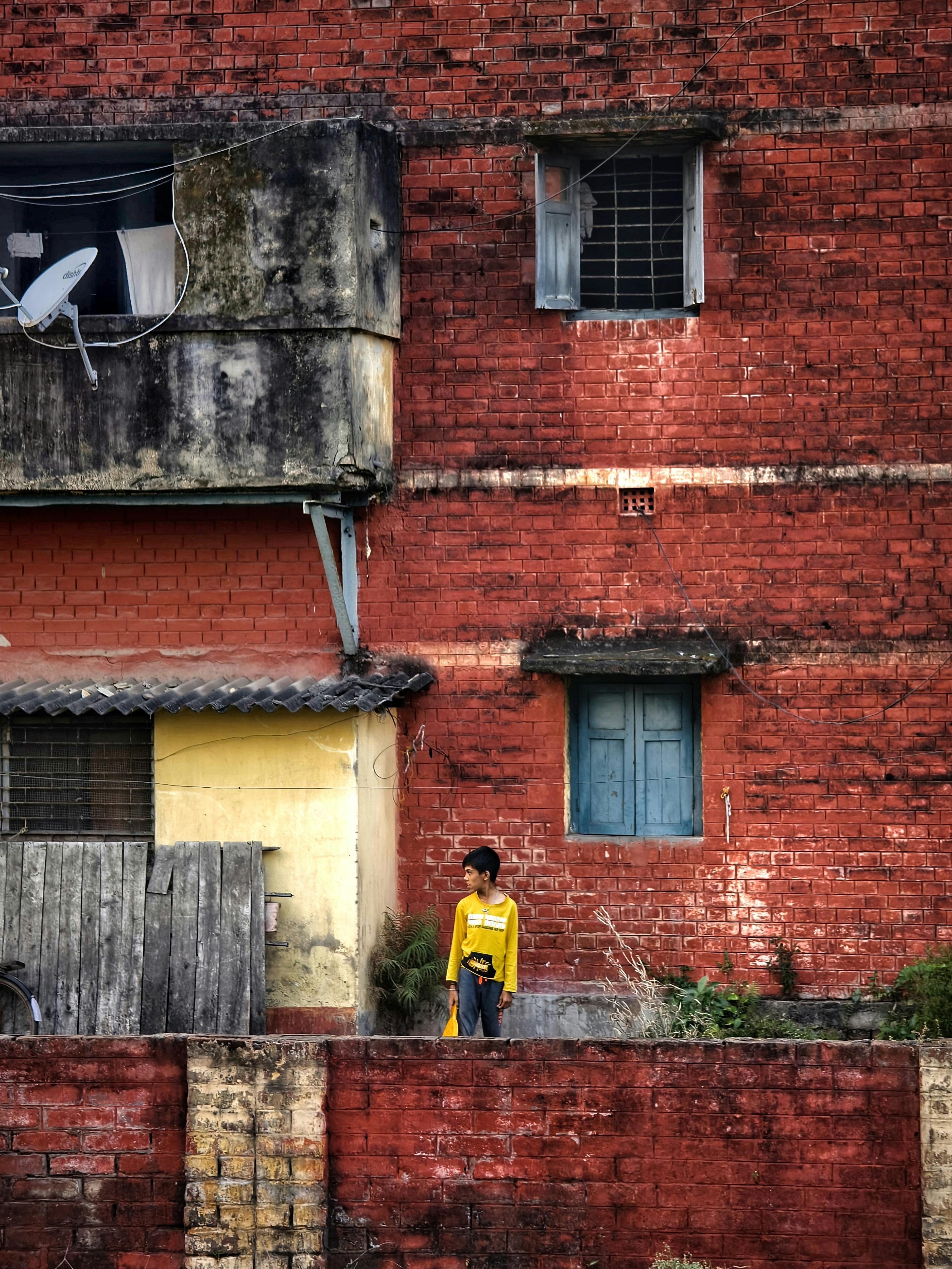 gratis Een kind in een geel shirt staat naast een felrode bakstenen muur in een landelijke, stedelijke omgeving. Stockfoto