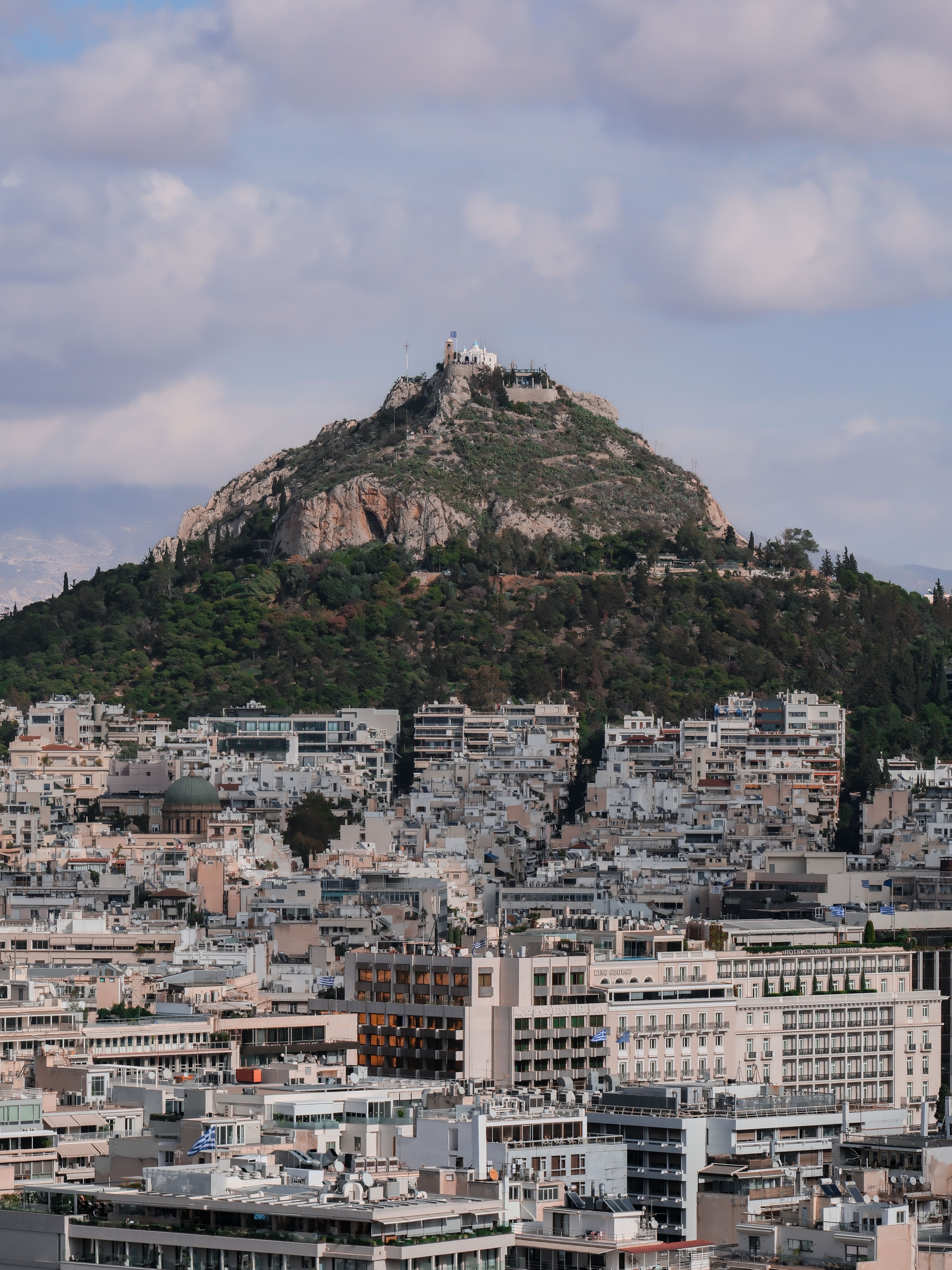 Aerial view of Mount Lycabettus overlooking Athens cityscape under a cloudy sky.