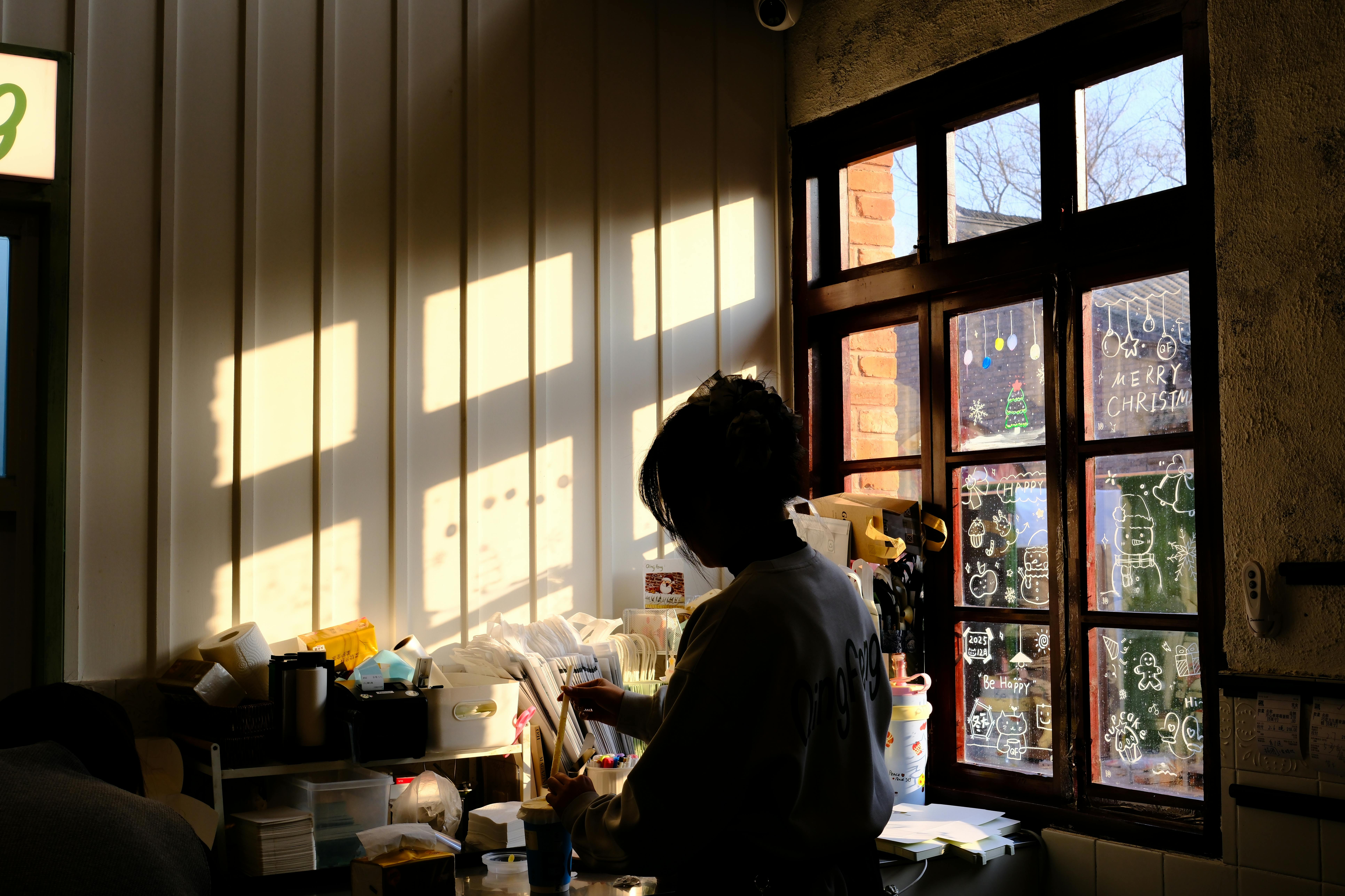 grátis Silhueta de uma pessoa trabalhando em um estúdio aconchegante, com arte festiva na janela e luz solar quente projetando sombras. Foto profissional