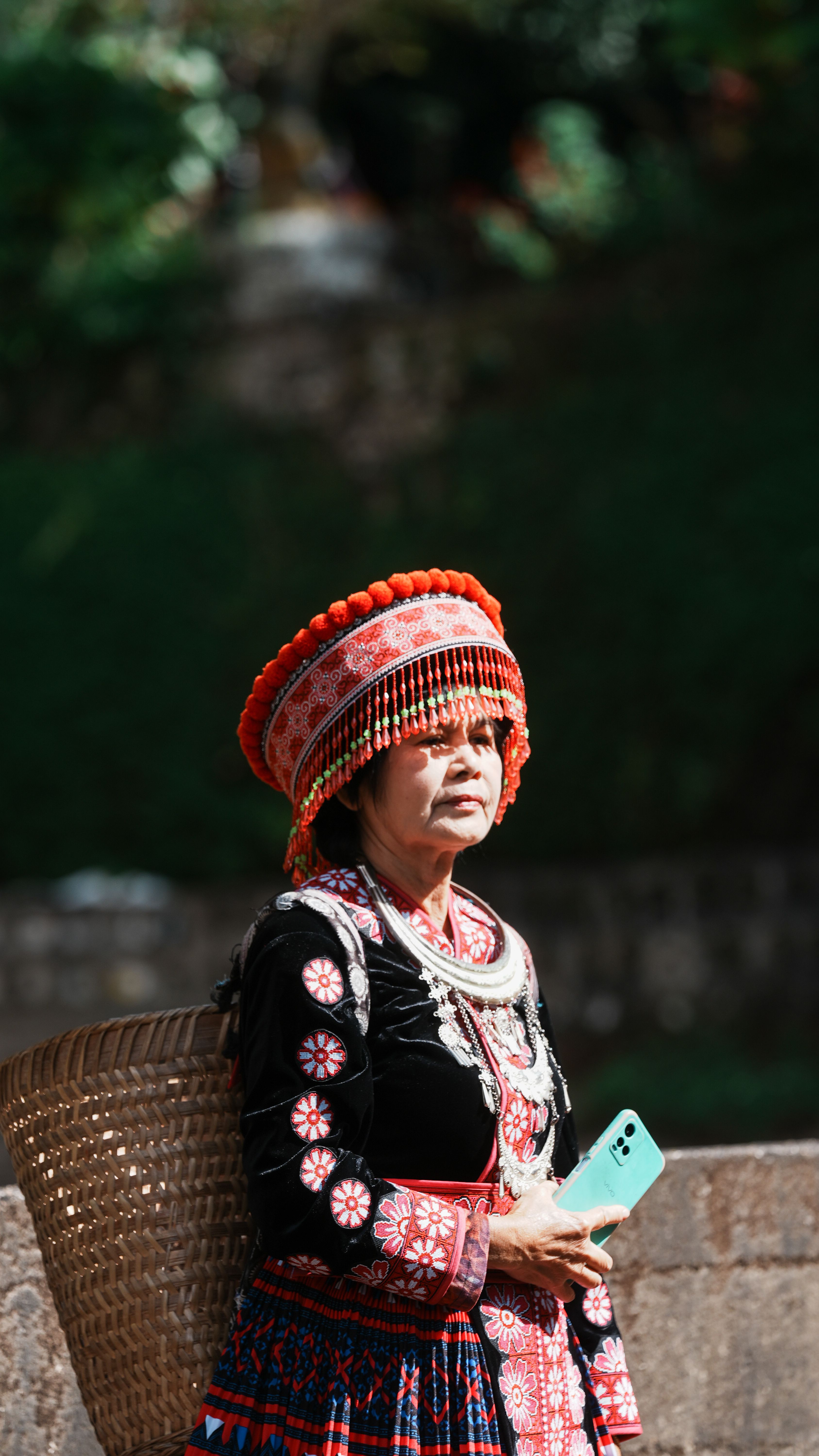 Woman in colorful traditional dress holding a phone and basket outdoors.