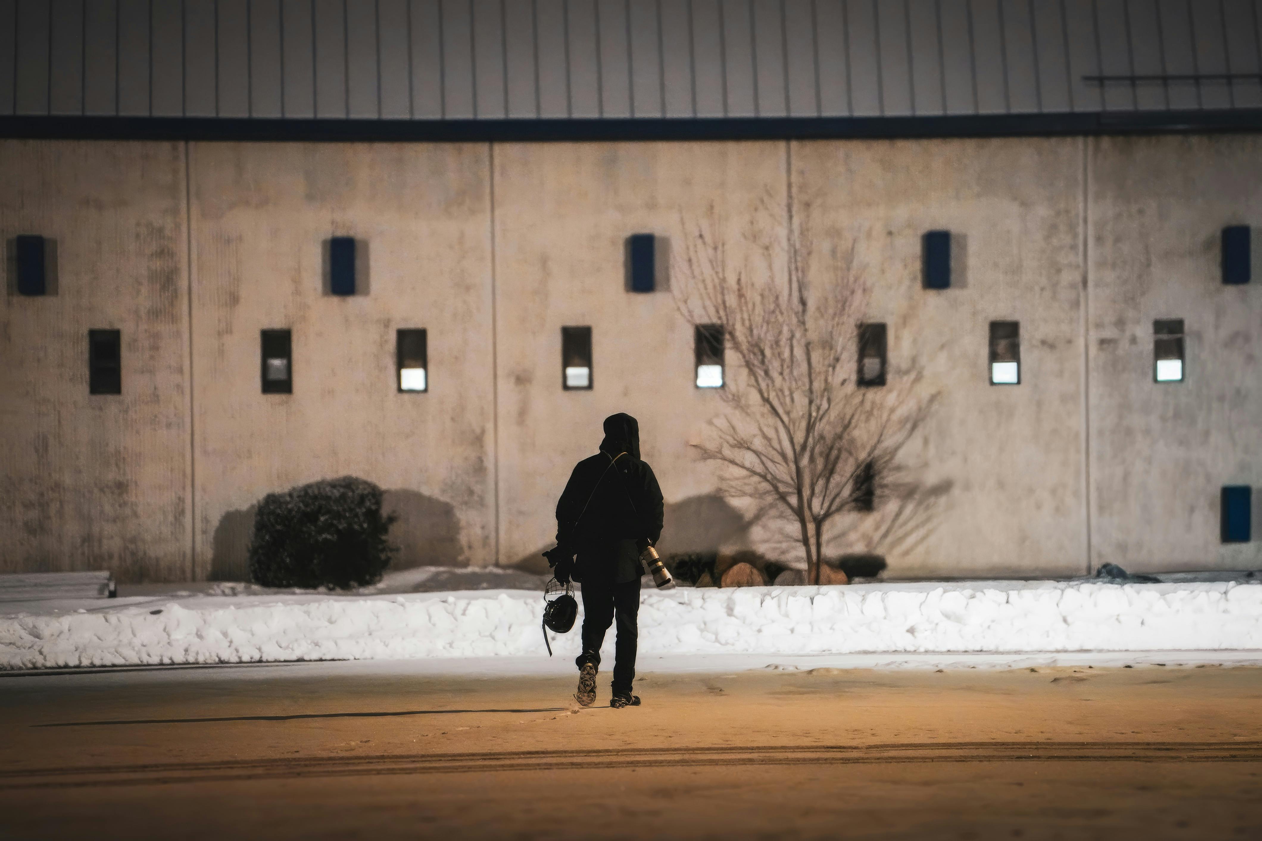 Δωρεάν Ένα άτομο περπατά μόνο του σε έναν χιονισμένο δρόμο τη νύχτα, κρατώντας μια φωτογραφική μηχανή, με φόντο ένα βιομηχανικό κτίριο. Φωτογραφία από στοκ φωτογραφιών
