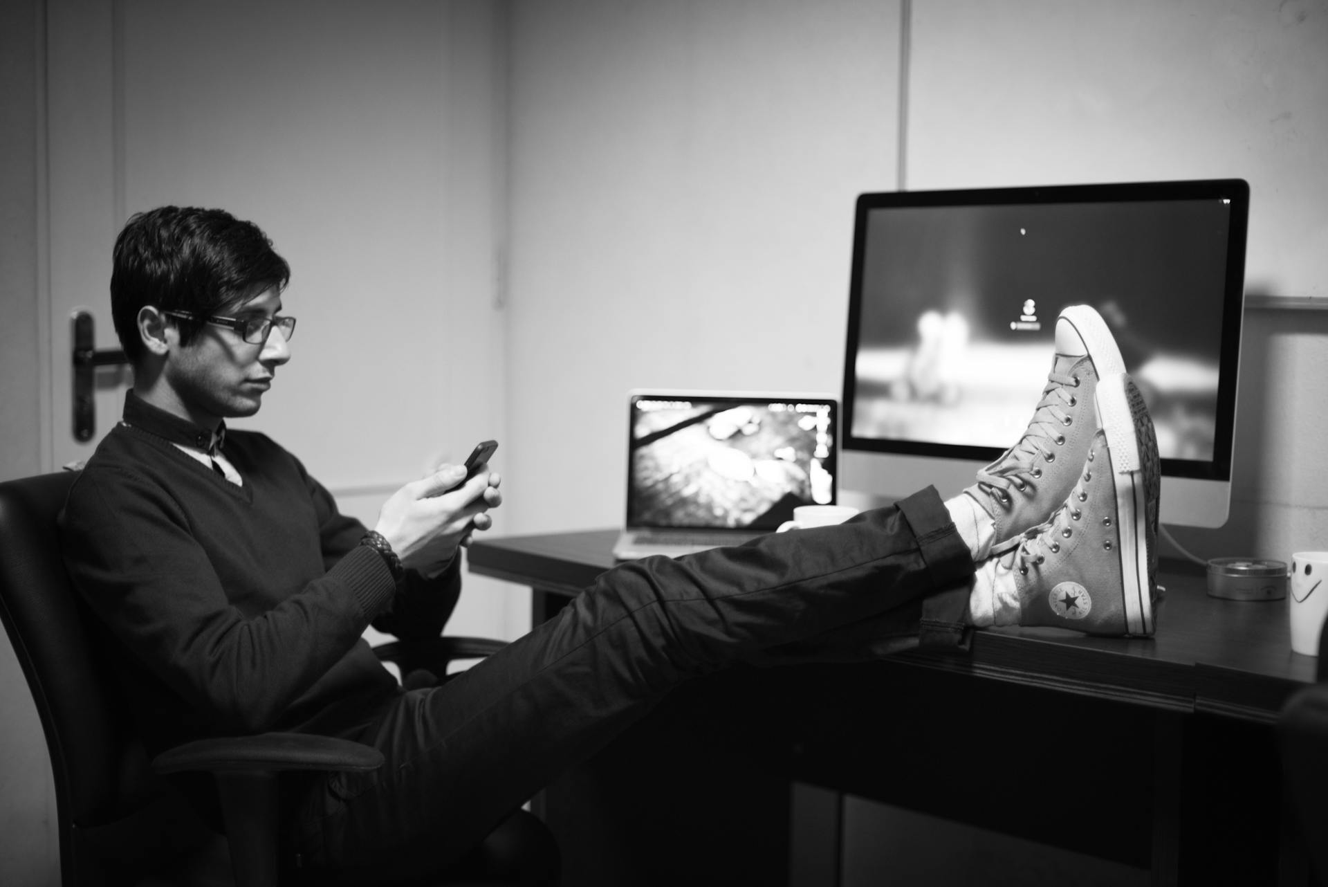 Man relaxing at desk using smartphone with feet up, featuring desktop and laptop computers.