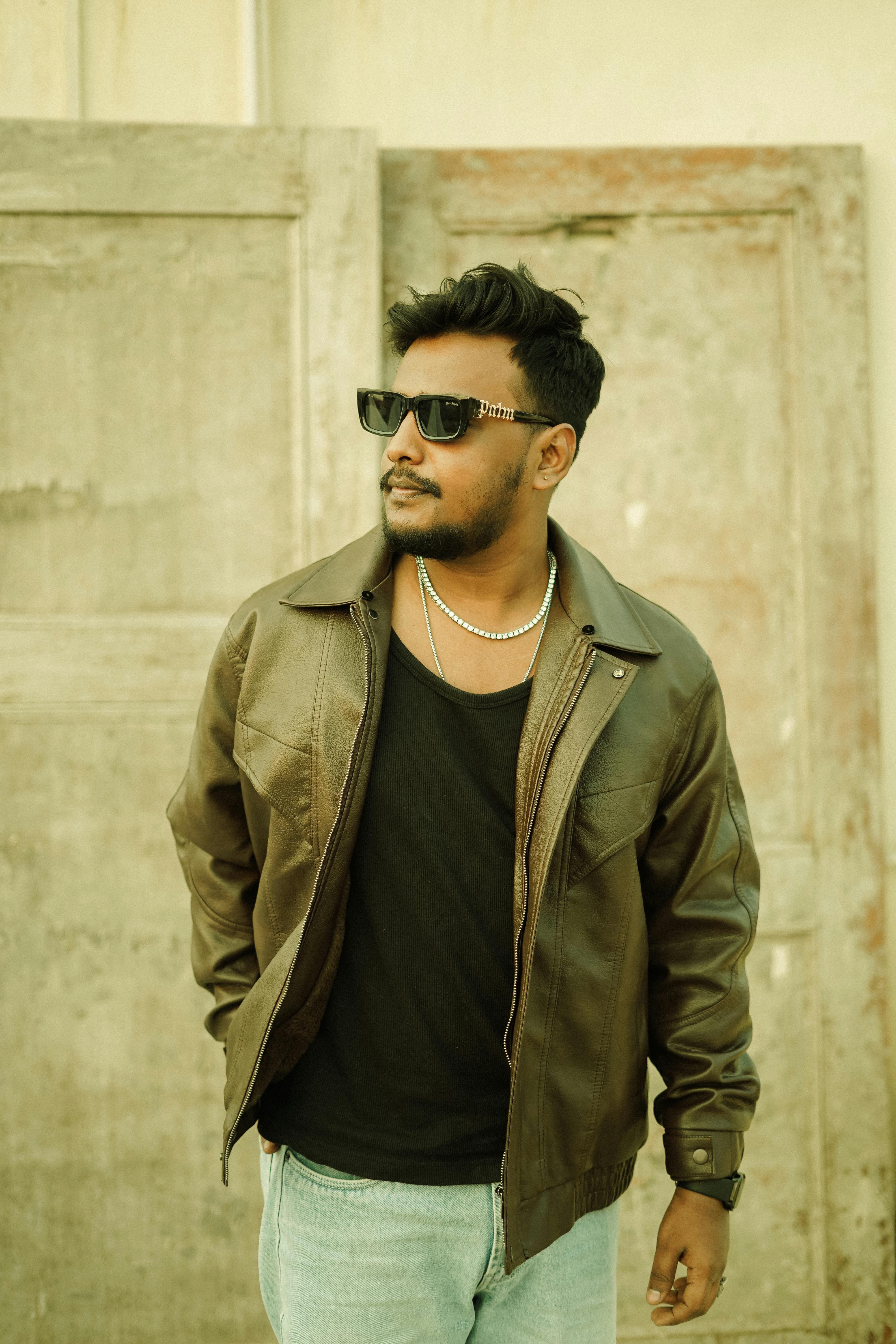Confident man in sunglasses and leather jacket poses against rustic background. Urban fashion scene.