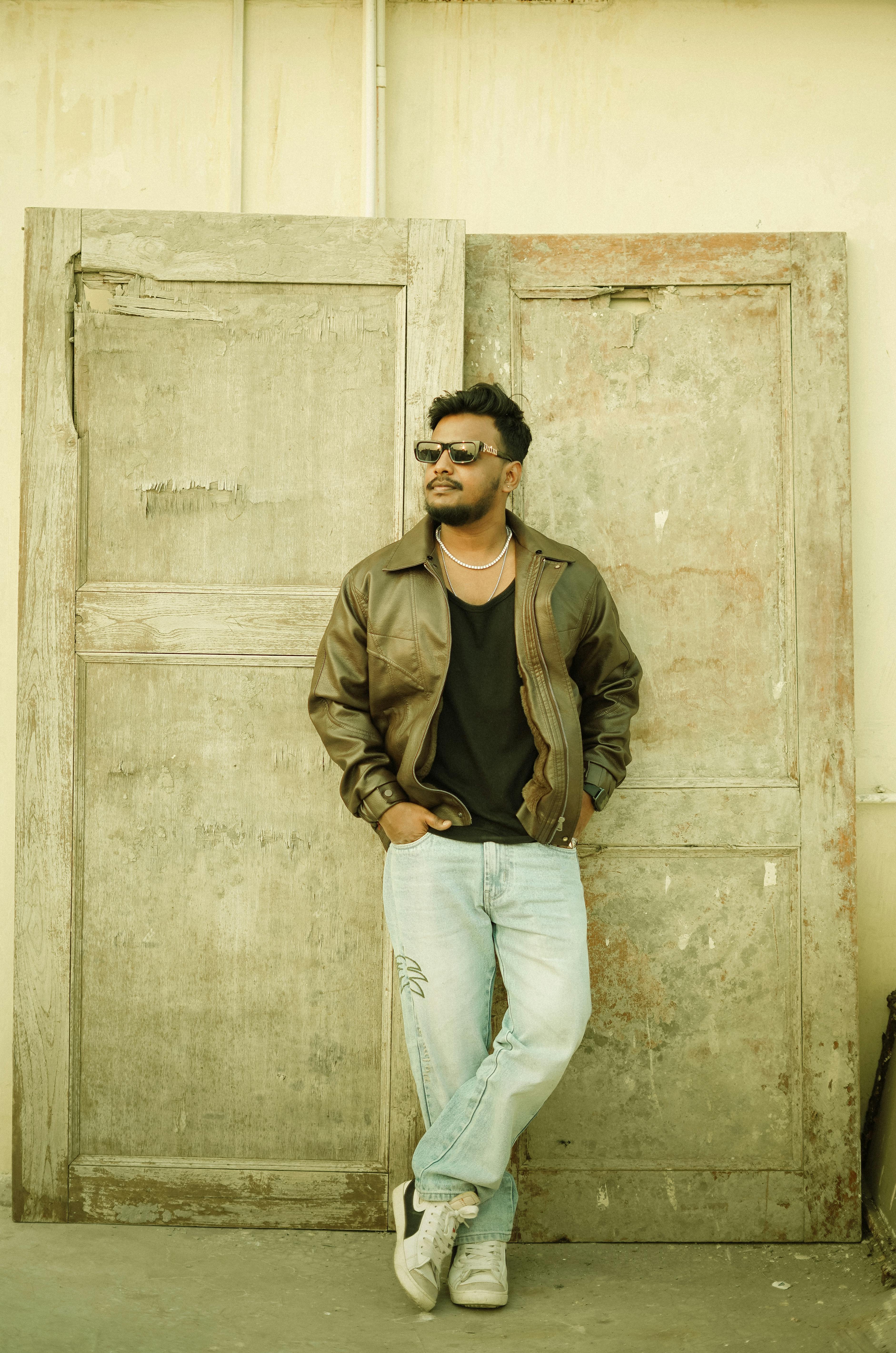 Man in leather jacket and sunglasses leaning against rustic doors.