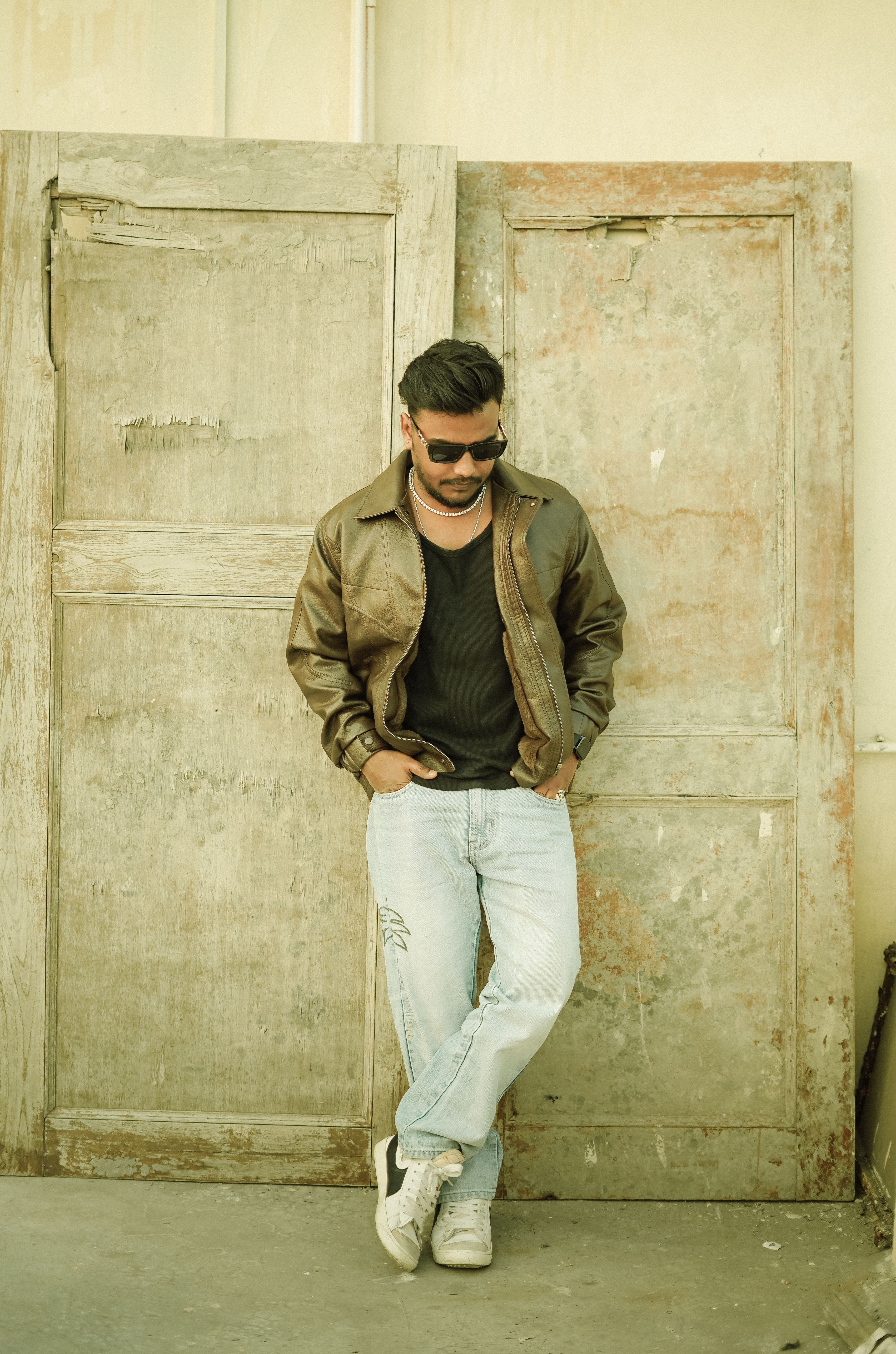 Stylish man wearing sunglasses and leather jacket posing casually against an old wooden door.
