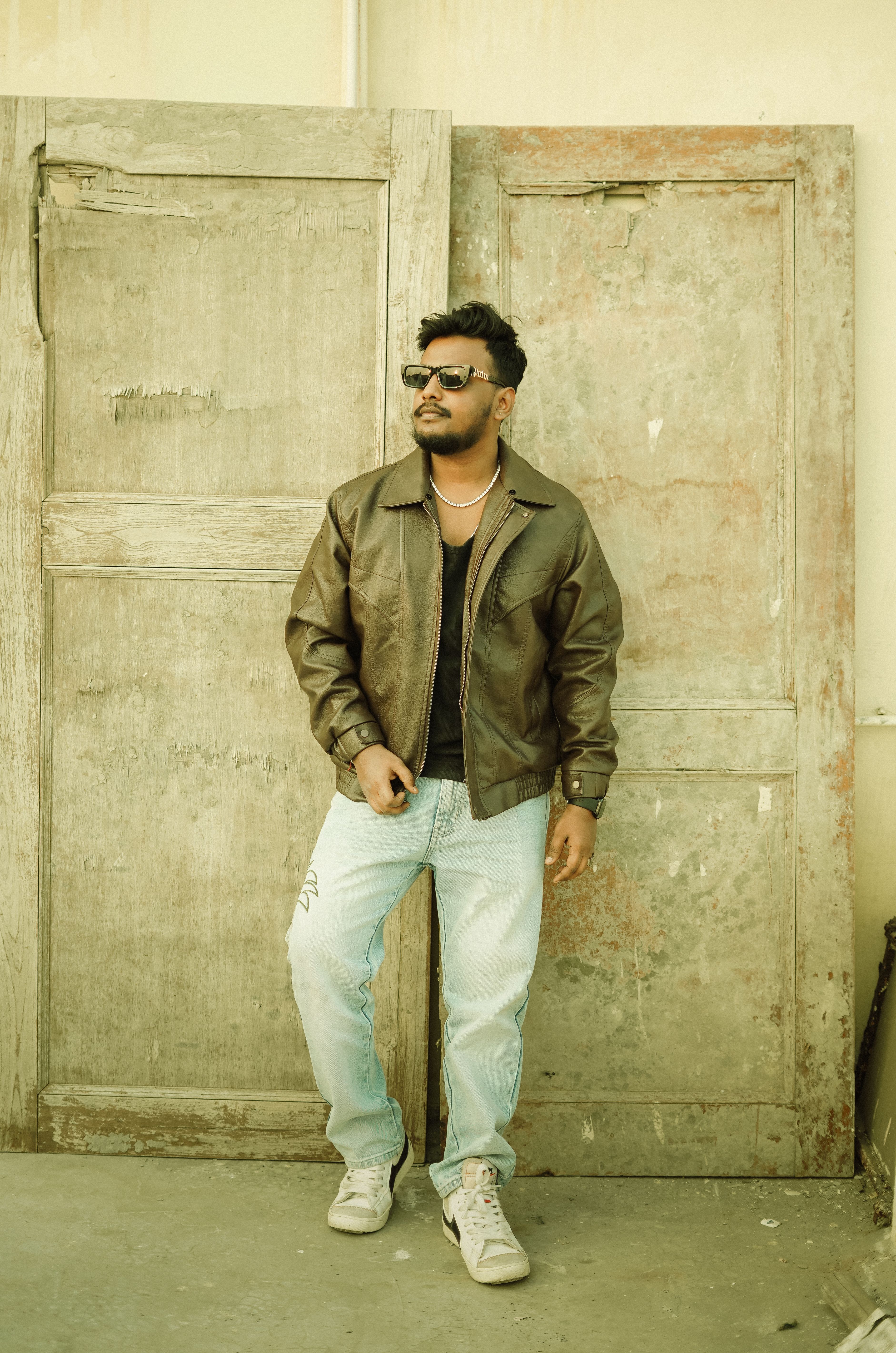 Fashion-forward man in a leather jacket and jeans poses against aged wooden doors. Urban style.