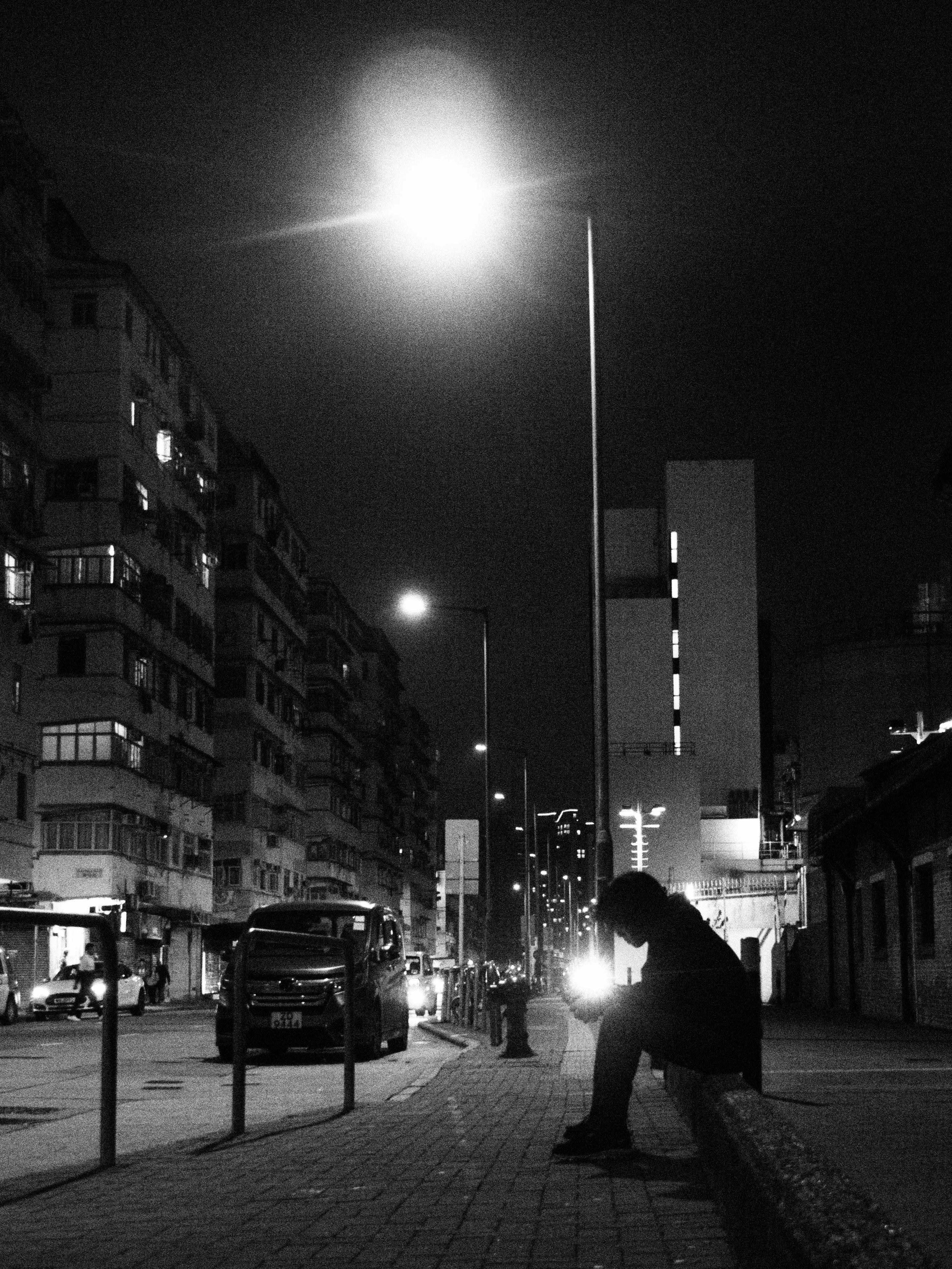 Gratis Svart-hvitt-bilde av en enslig skikkelse som sitter under gatelykter på en bygate om natten. Arkivbilde