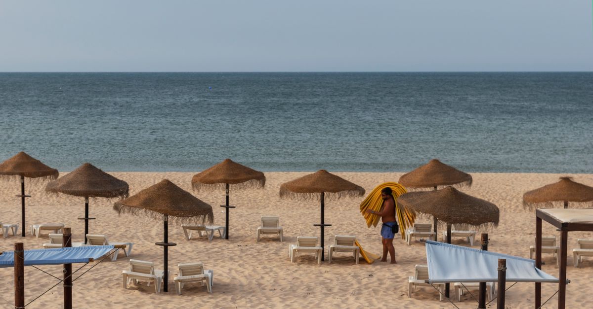Uma cena de praia tranquila com guarda-sóis de palha e uma pessoa preparando espreguiçadeiras à beira-mar.