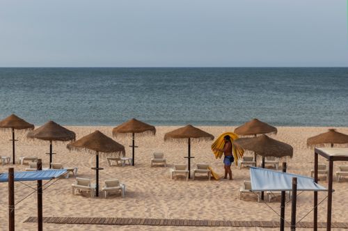 Free A tranquil beach scene with straw umbrellas and a person preparing sun loungers by the sea. Stock Photo