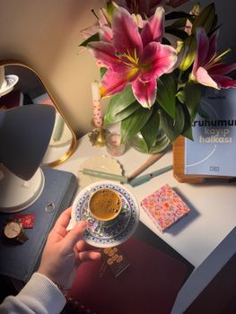 A cozy desk with coffee, vibrant pink lilies, and books, perfect for relaxation.