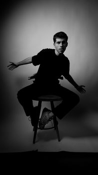 Expressive black and white portrait of a young man posed dynamically on a stool in a photo studio.