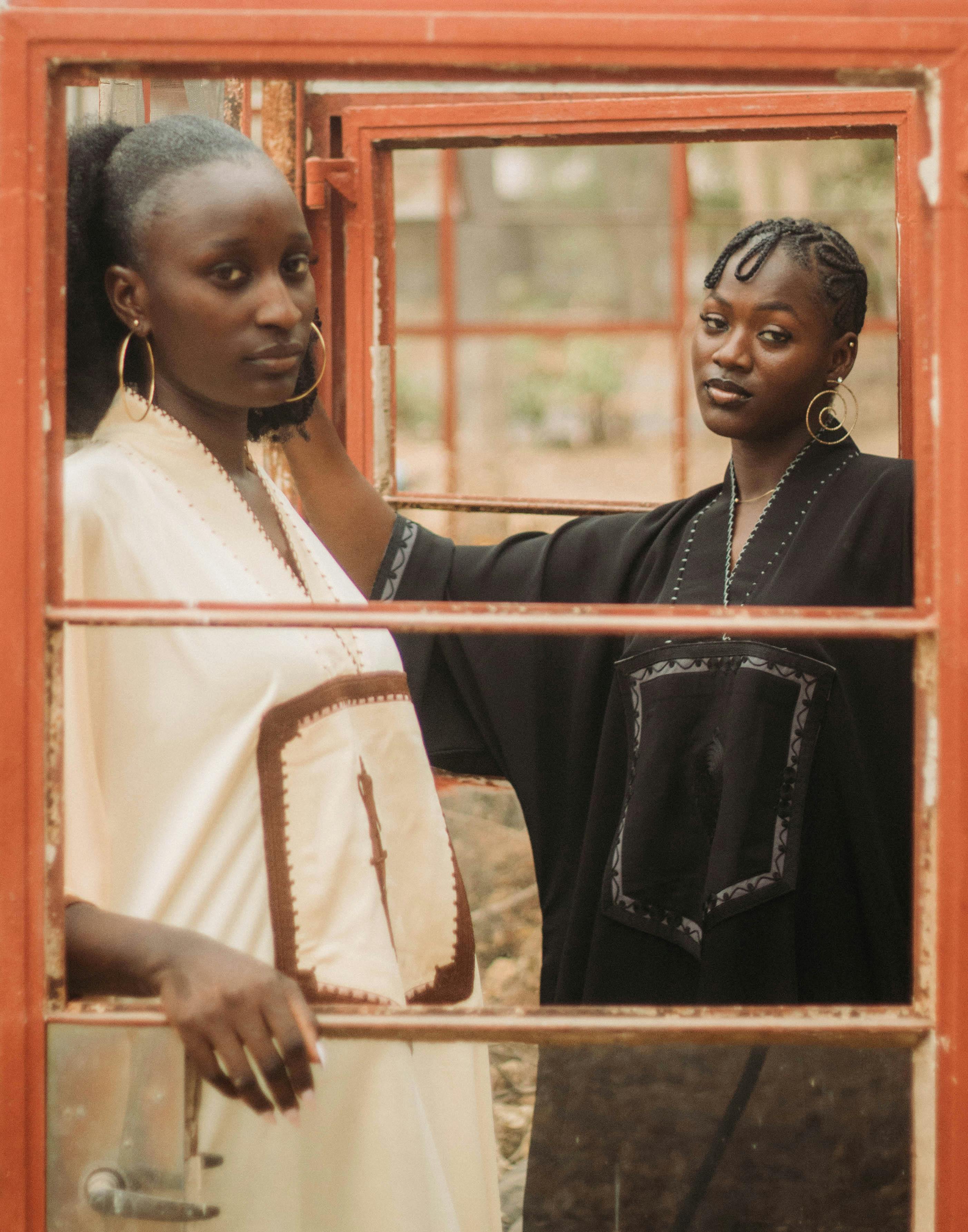 grátis Duas mulheres com trajes elegantes, fotografadas através de uma moldura de janela antiga, exalam elegância e graça. Foto profissional