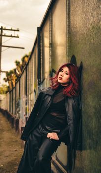 Fashionable woman with red hair in a leather outfit poses against a graffiti-covered wall in an urban alley.