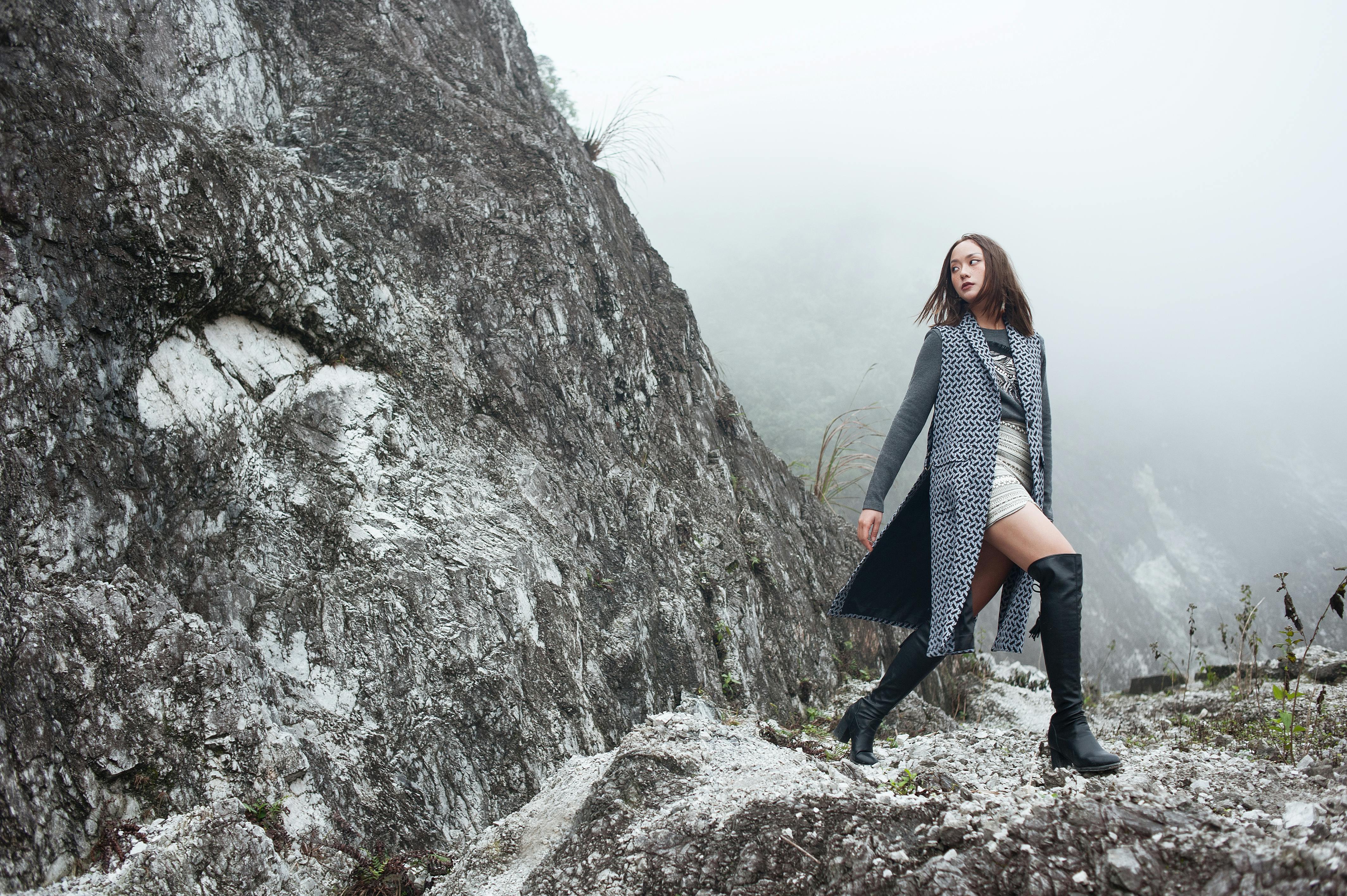 Fashionable woman in boots poses confidently on a foggy rocky cliff, showcasing modern style amidst rugged nature.