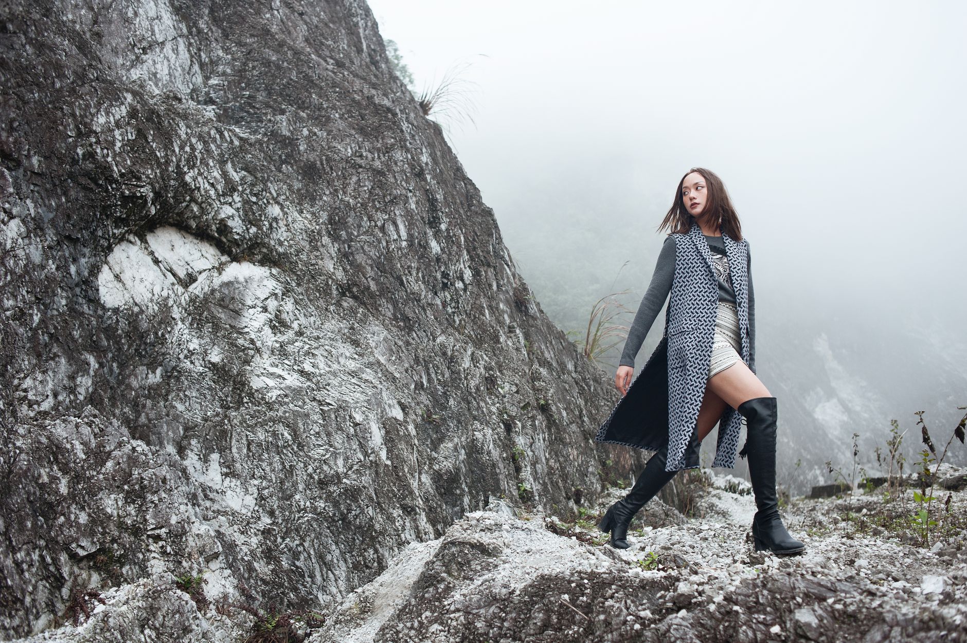 Fashionable woman in boots poses confidently on a foggy rocky cliff, showcasing modern style amidst rugged nature.