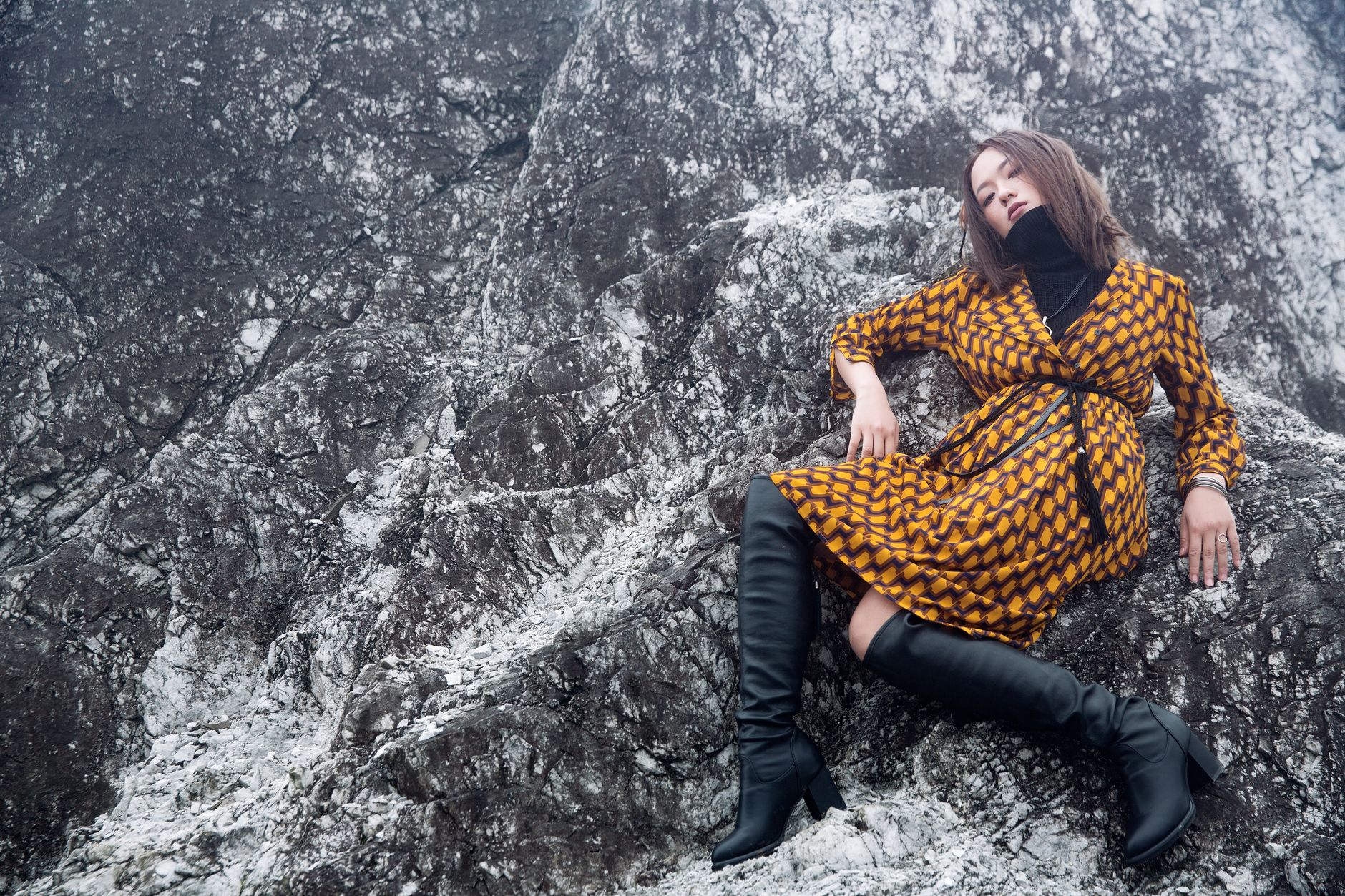 A stylish woman in a patterned dress and boots poses against rugged rocks outdoors.