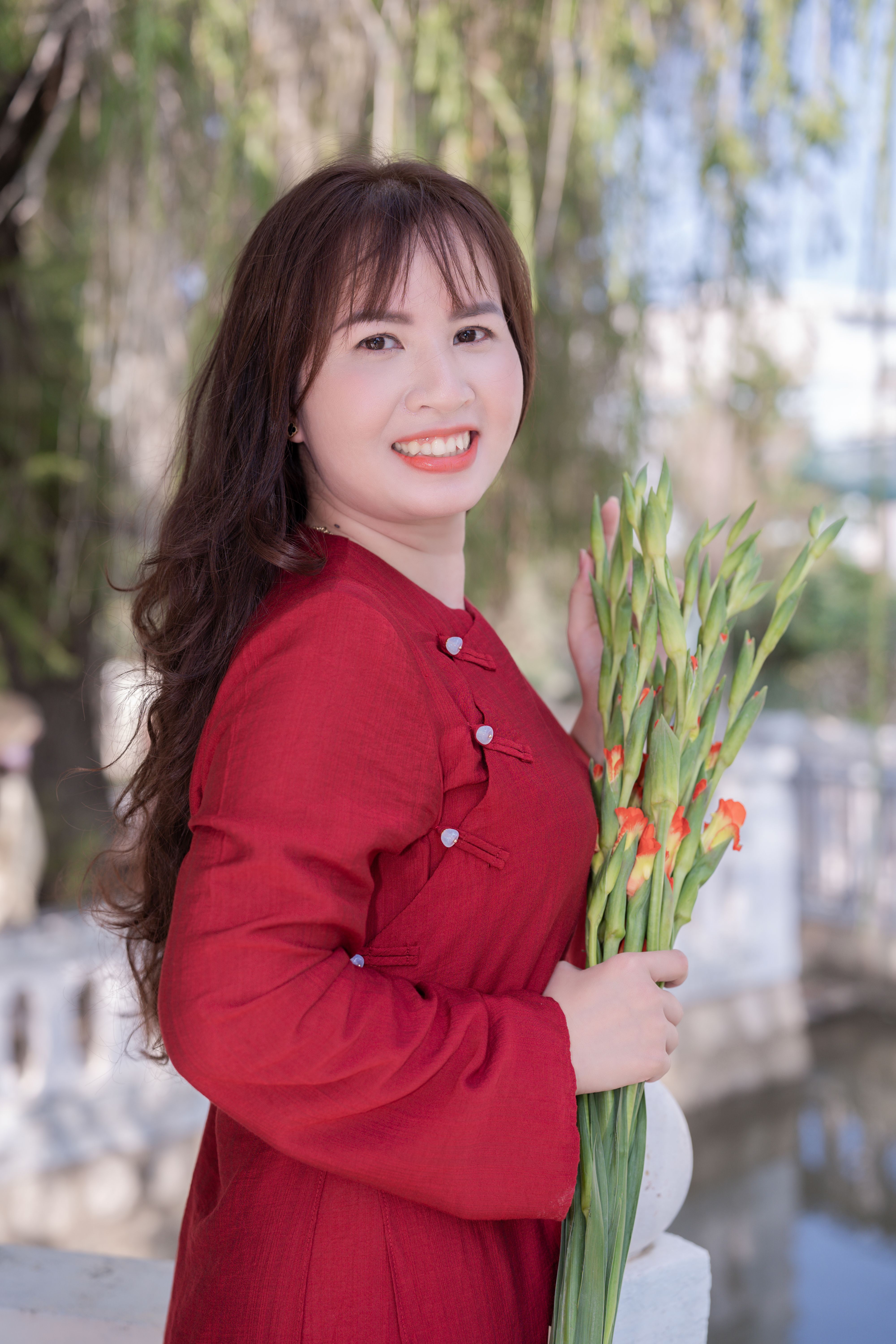 Smiling woman in a red dress holding gladiolus flowers outdoors.