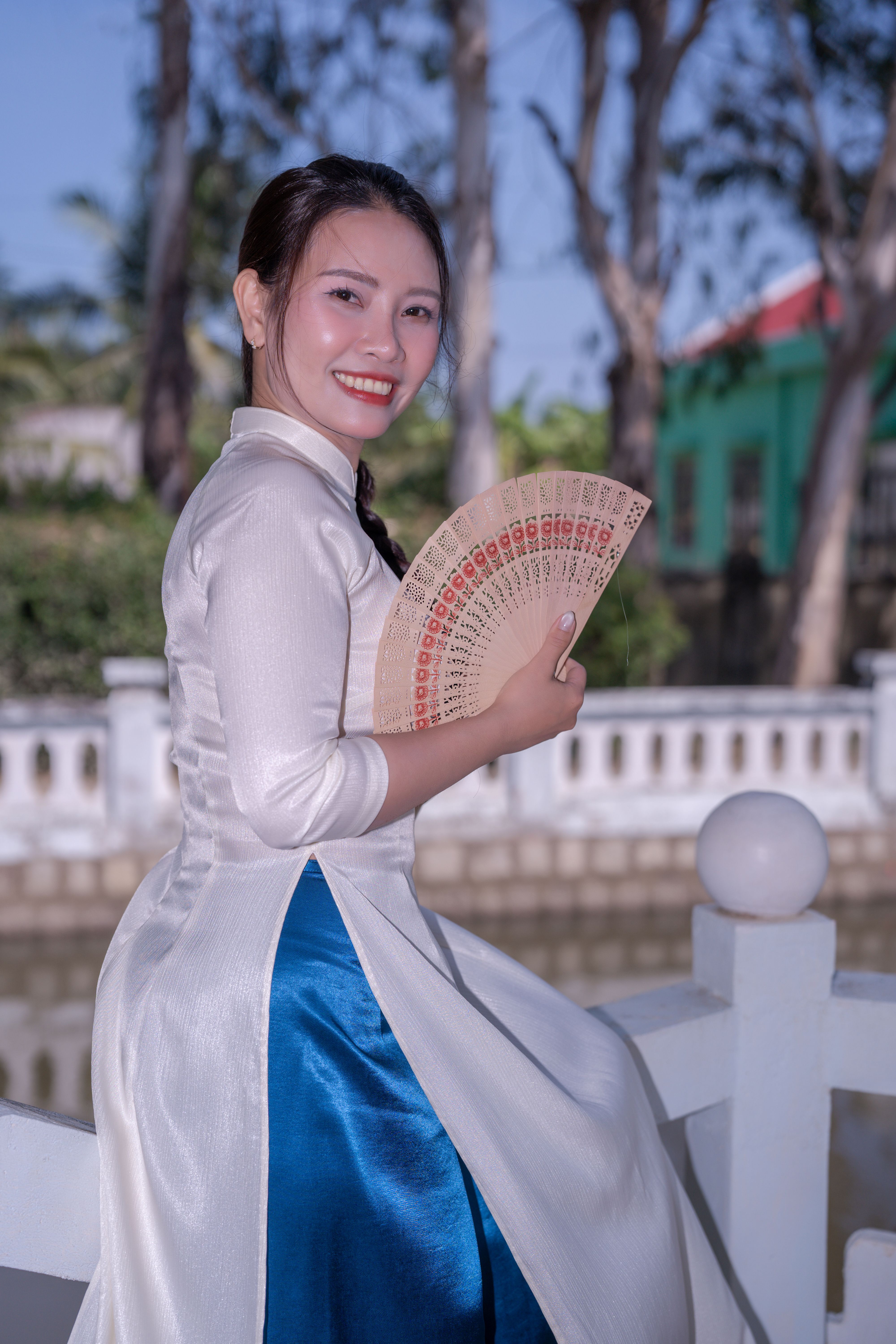Smiling woman in traditional Vietnamese Ao Dai holding a fan, outdoors with a serene background.