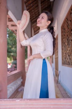 Woman in traditional white dress holding a fan, smiling elegantly.