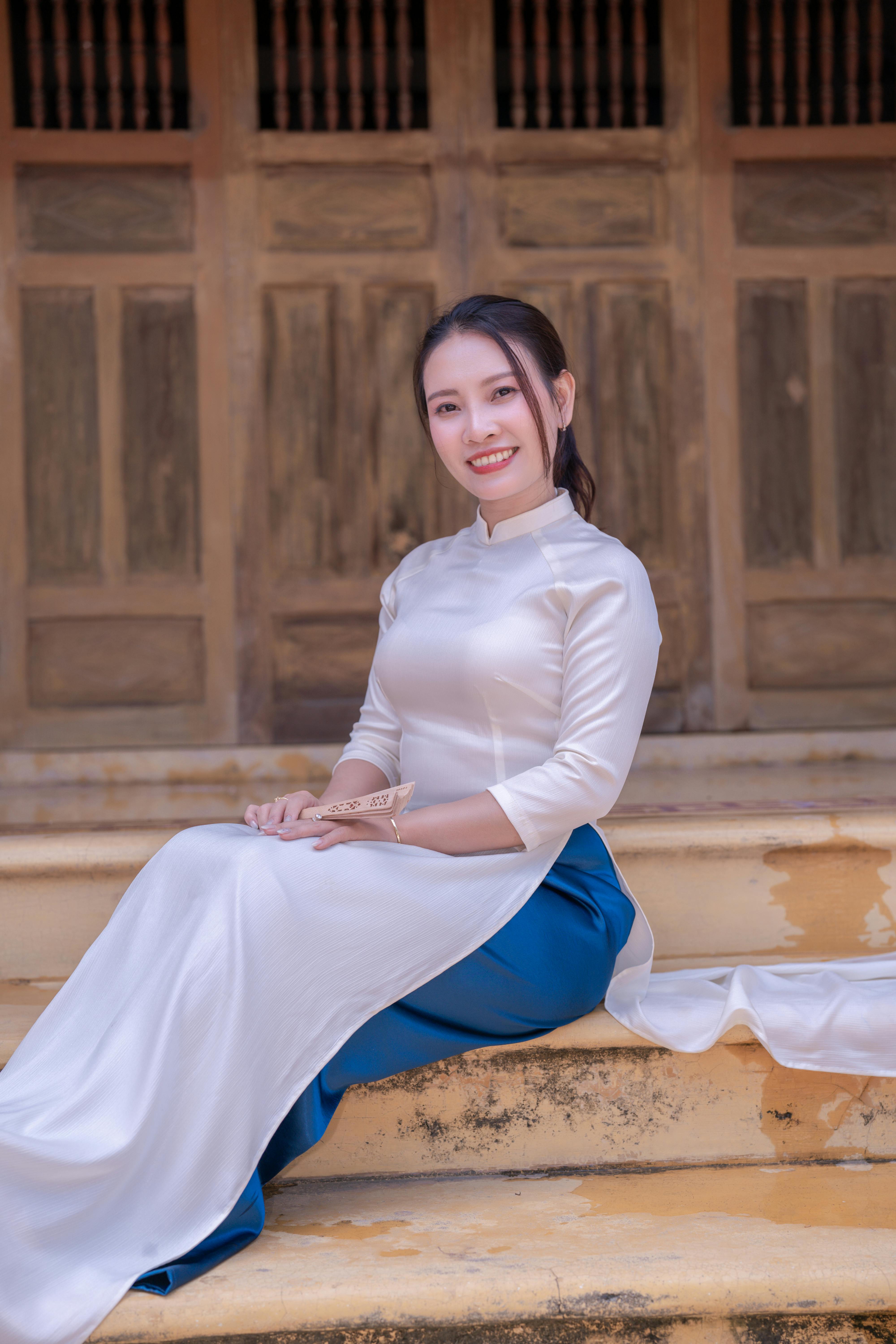 Smiling woman wearing traditional Vietnamese Ao Dai seated on steps with a wooden door backdrop.