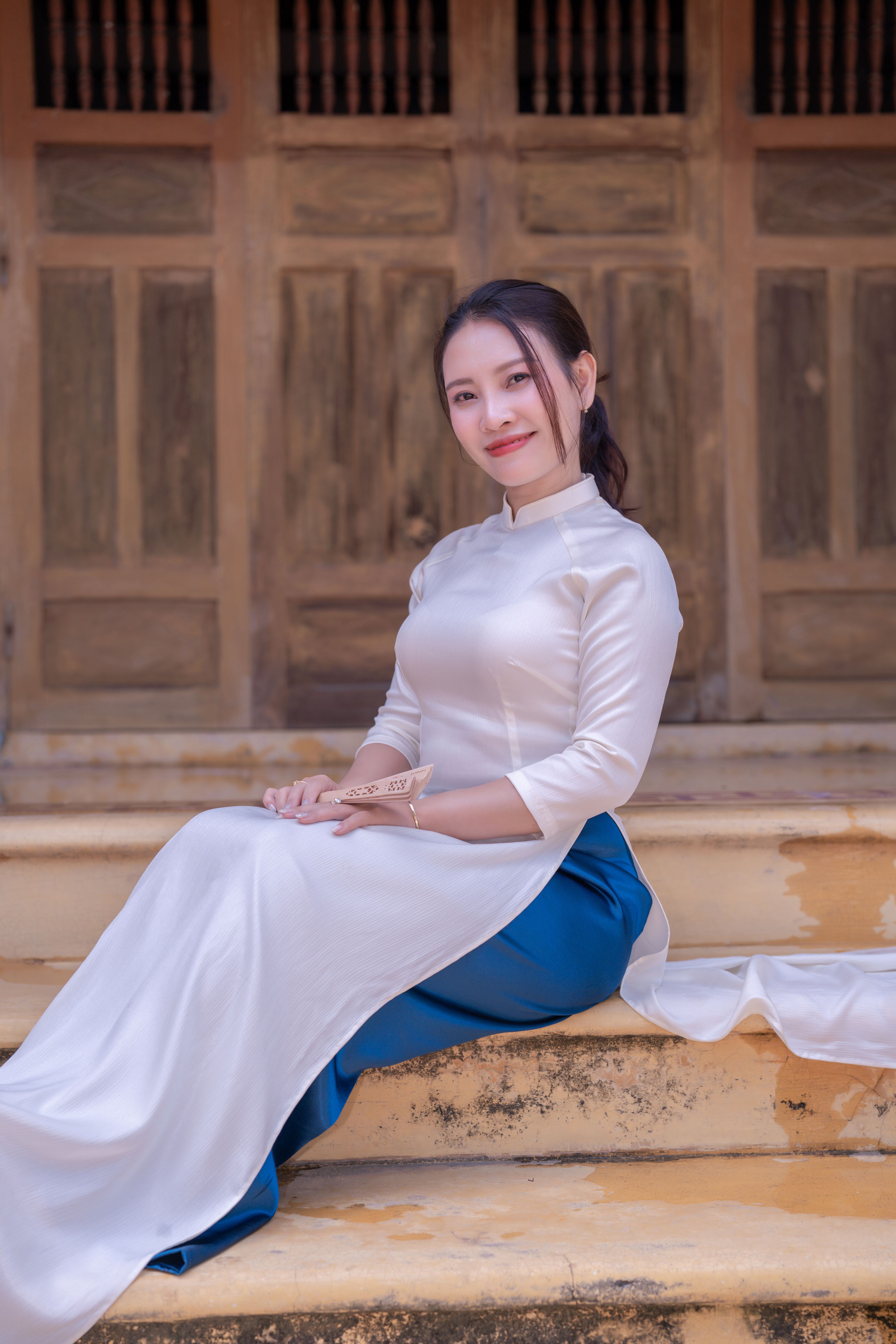 Smiling woman wearing traditional white Áo Dài sitting on steps with wooden background.