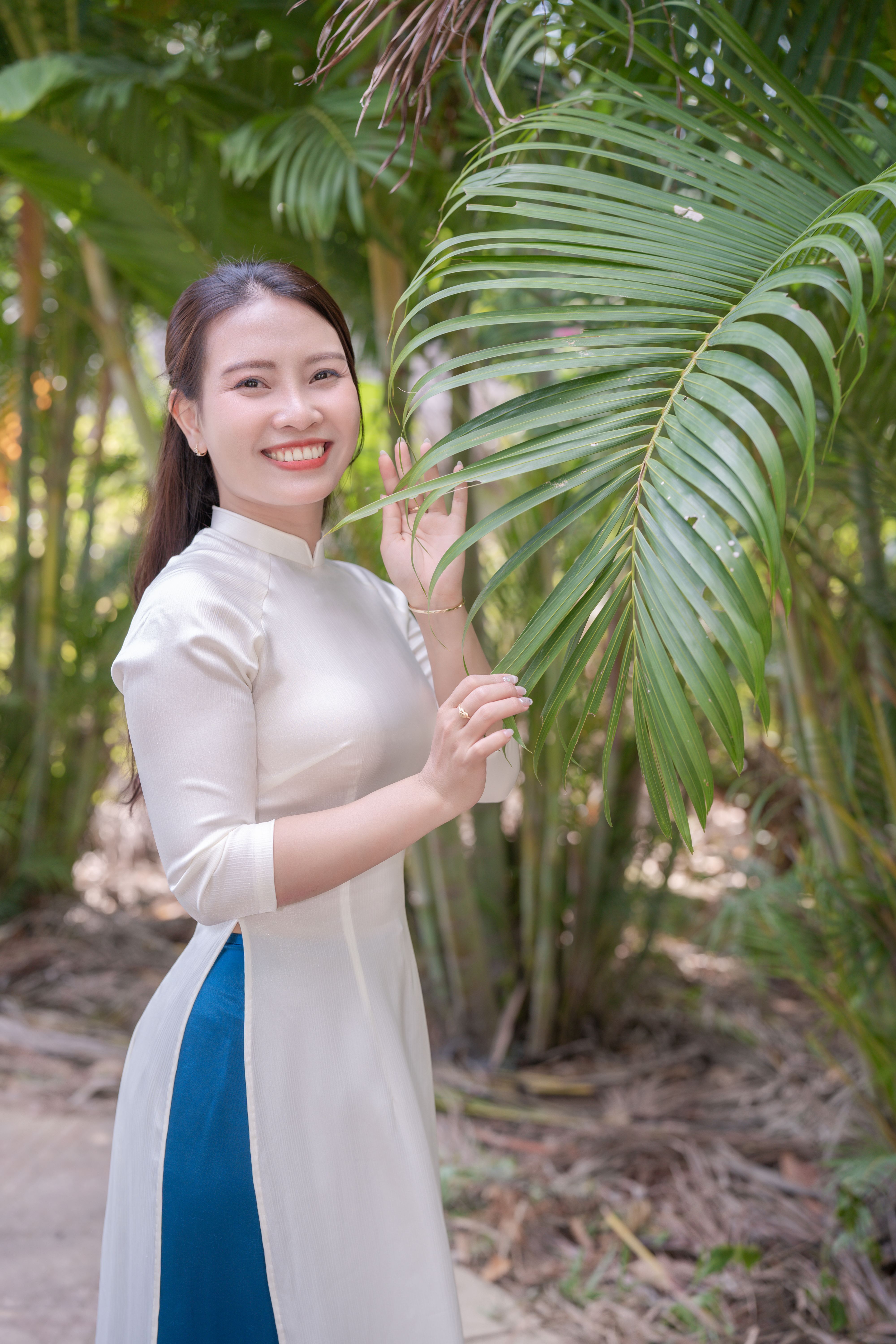 Young woman in white ao dai stands amidst greenery, smiling joyfully.