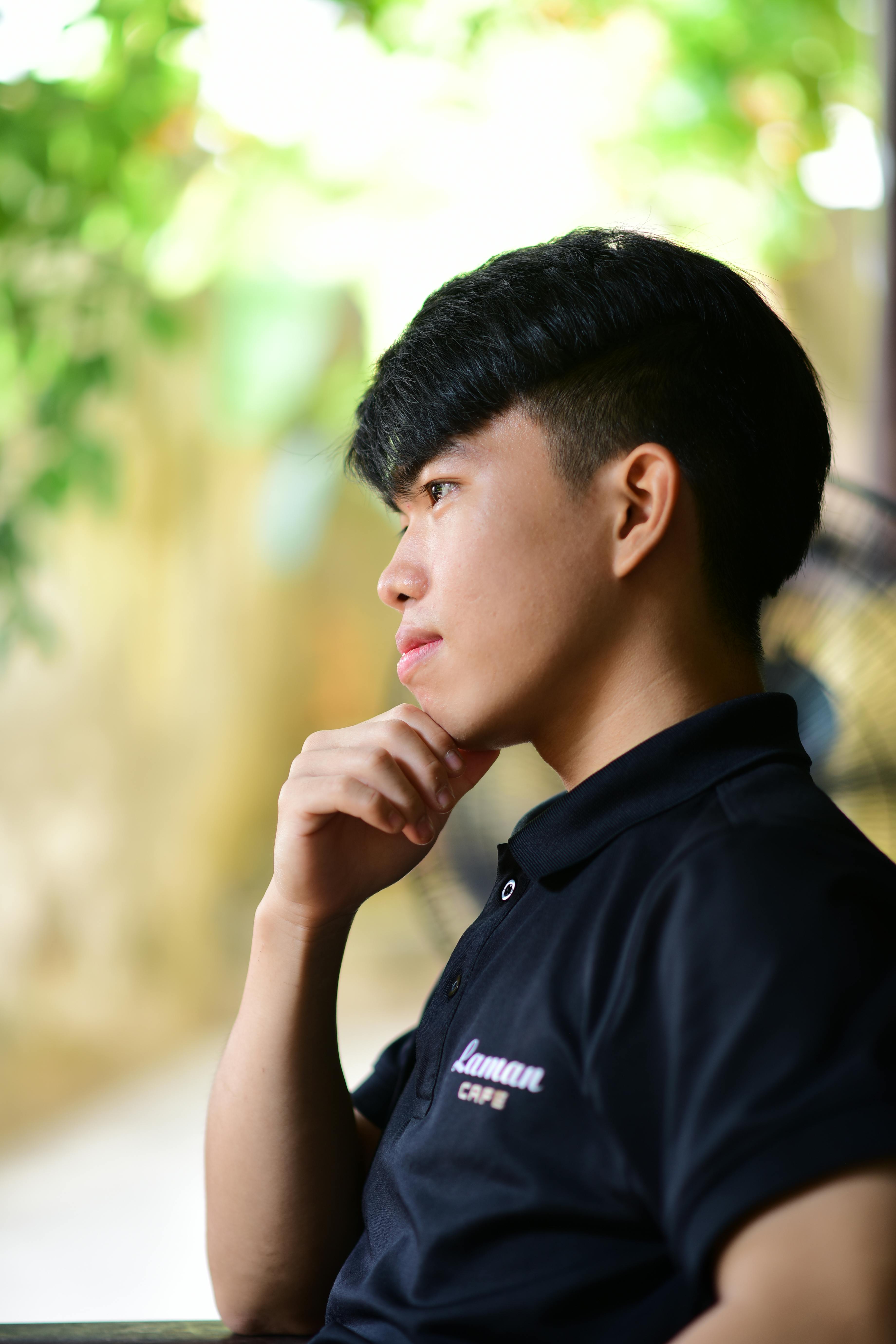 Free Profile view of a young man deep in thought, seated outdoors with blurred green background. Stock Photo