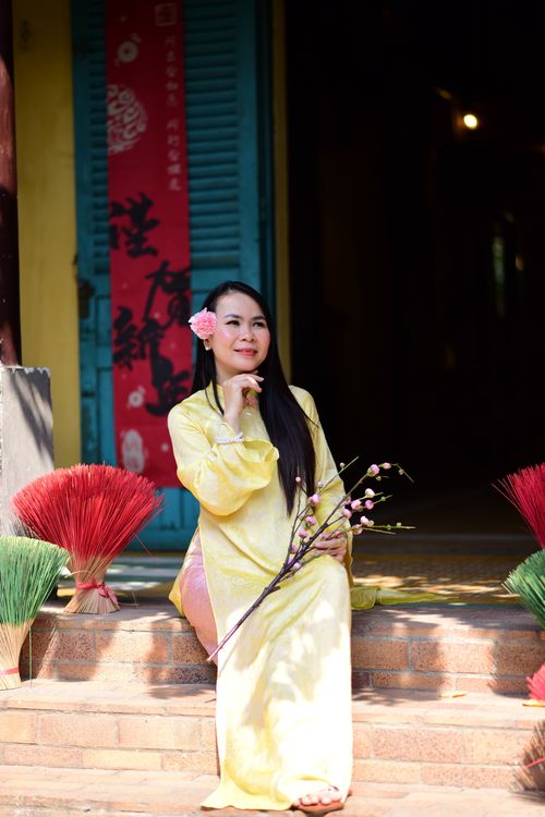 Free A woman in a yellow traditional dress sits on steps with pink flower bouquet, surrounded by colorful brooms. Stock Photo
