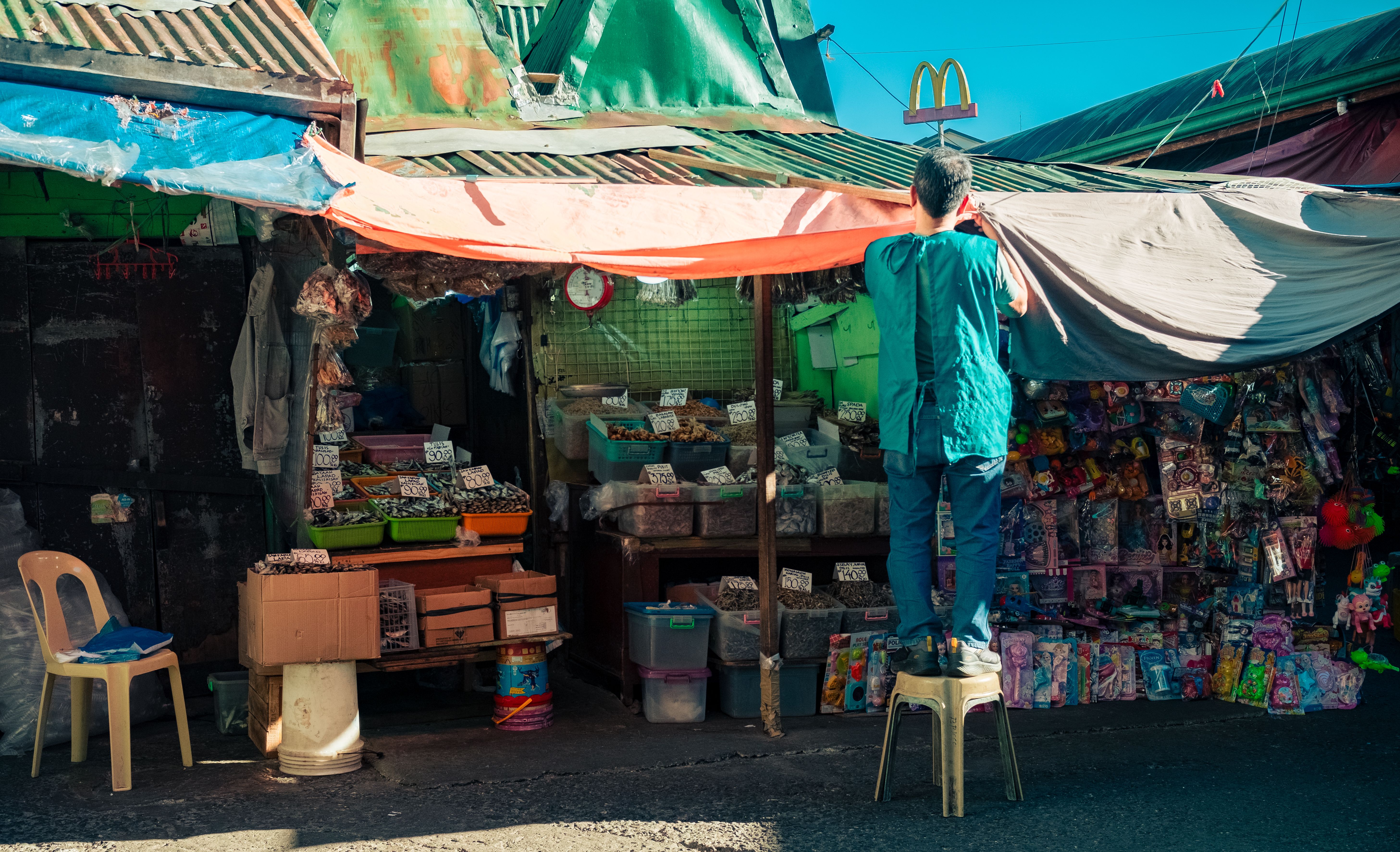 A vendor adjusts a tarp at a vibrant outdoor market stall overflowing with colorful products.