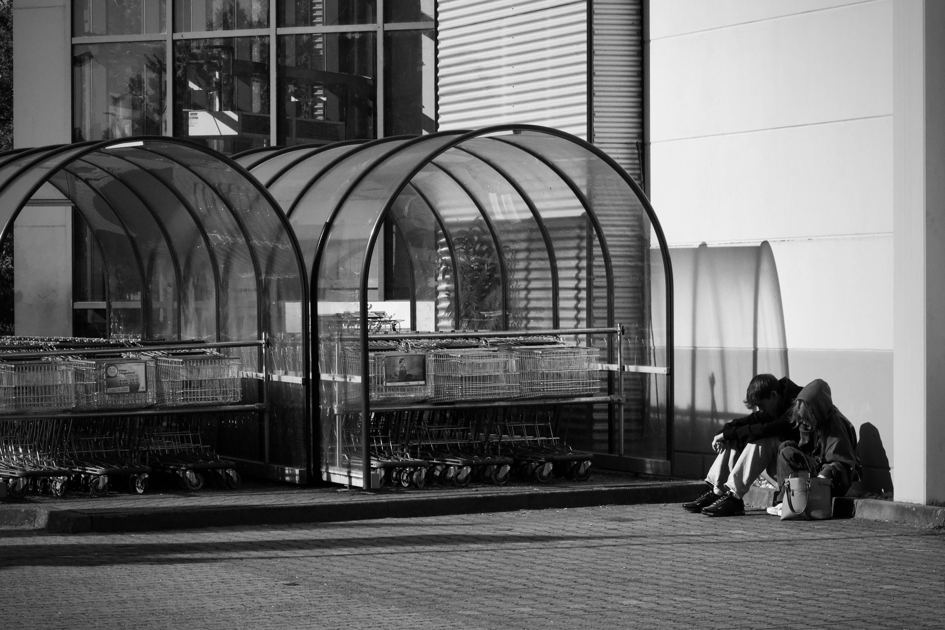 Gratis Imagen monocromática de dos personas sentadas cerca de refugios para carritos de compras en el exterior. Foto de stock