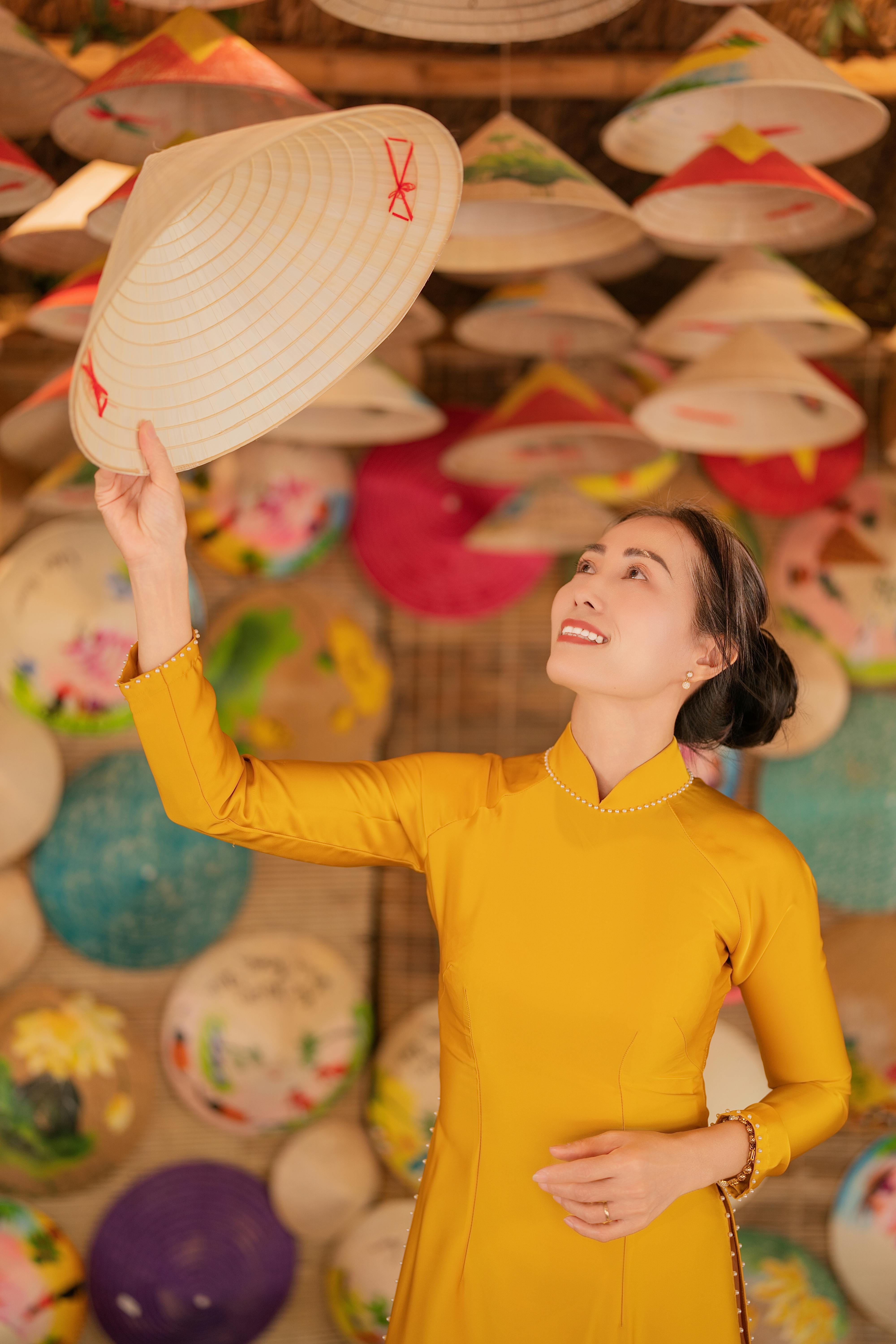 Woman in yellow ao dai holding a Vietnamese conical hat, smiling against a colorful backdrop of hats.