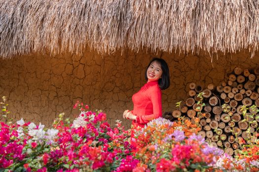 A woman in a vibrant red dress stands smiling among colorful flowers against a rustic backdrop.