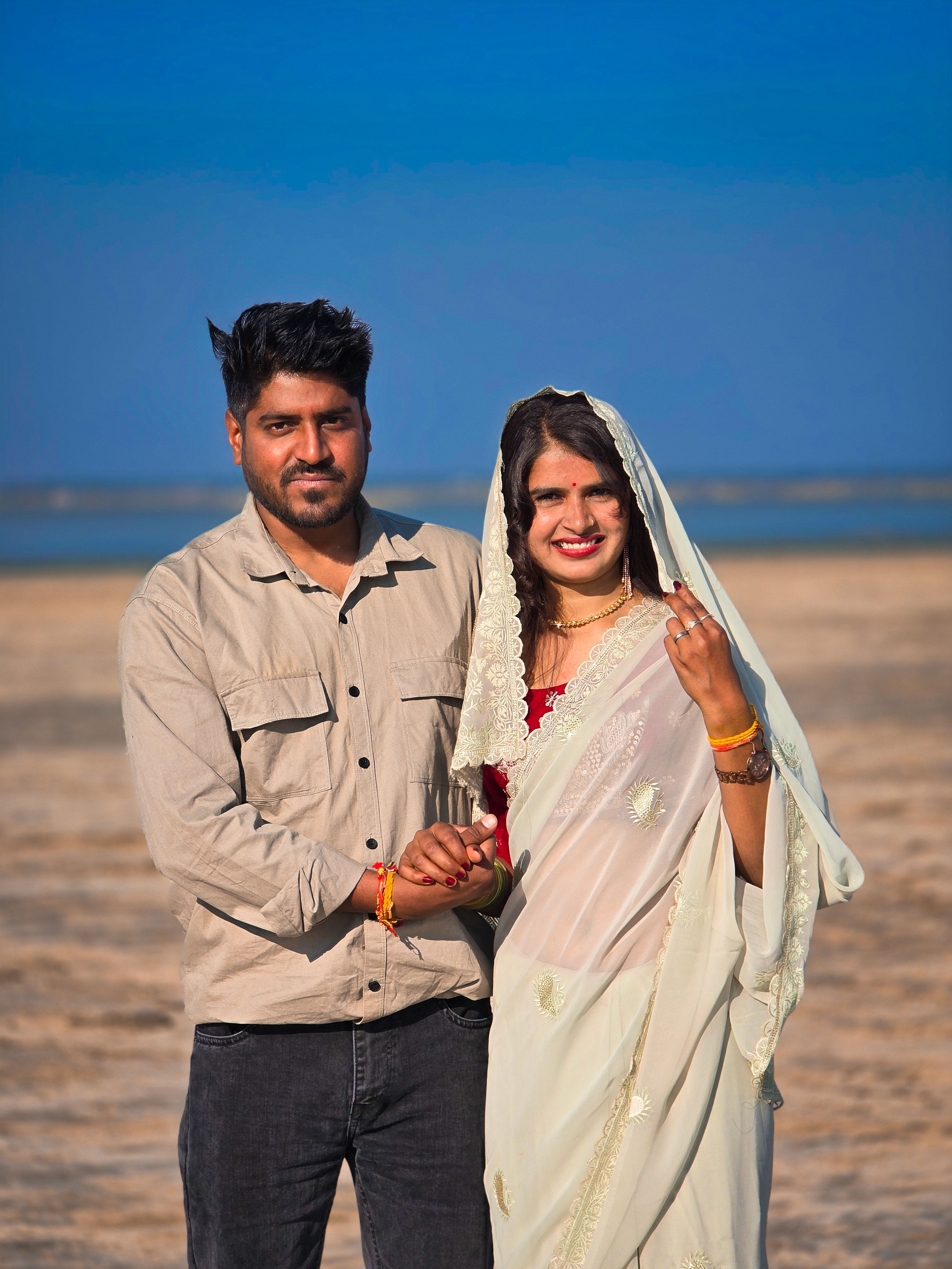 A joyful couple in traditional clothing posing on a sunny beach with a clear blue sky.