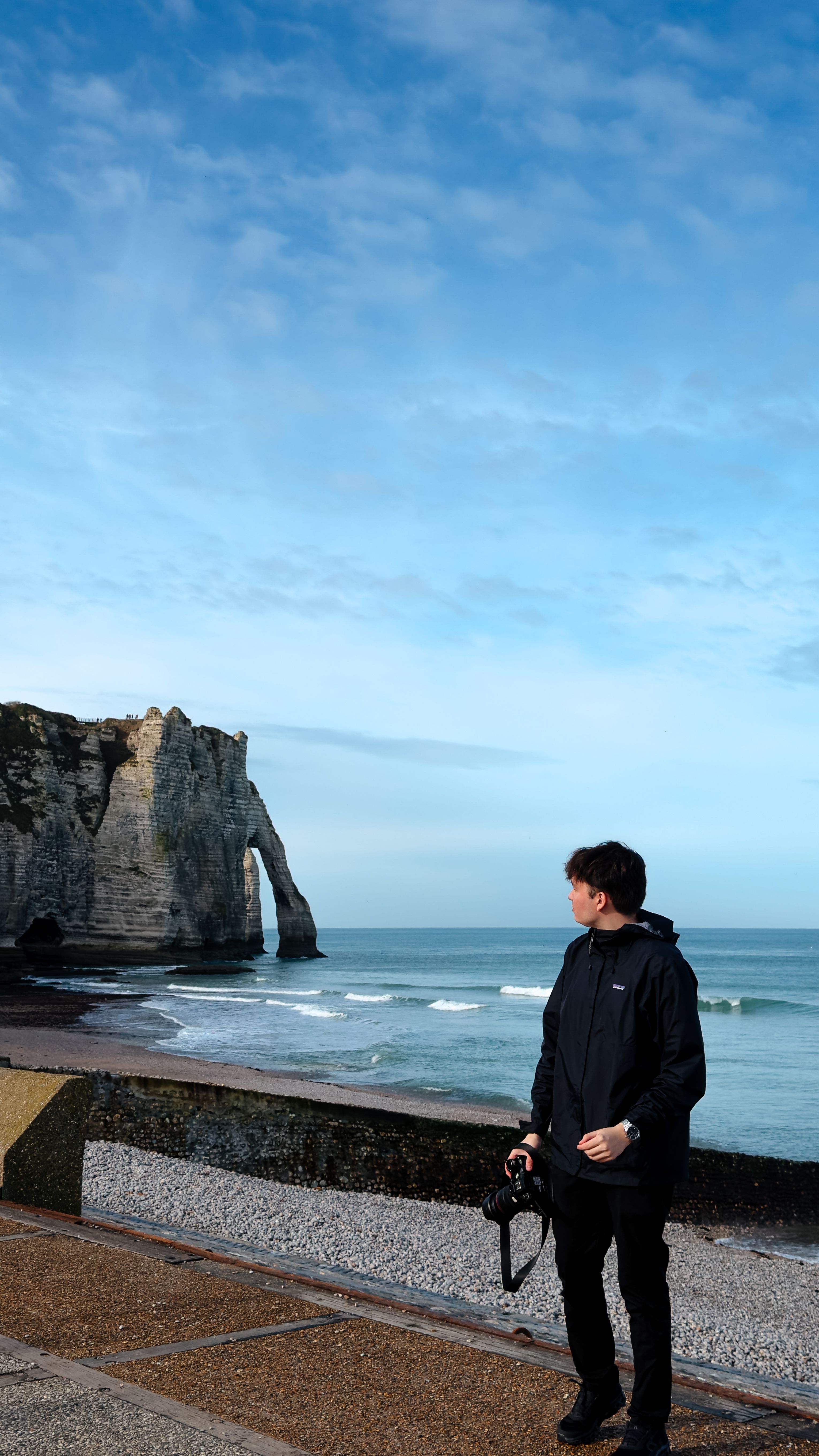 A photographer stands by the iconic cliffs of Étretat, Normandy, holding a camera, showcasing natural beauty.