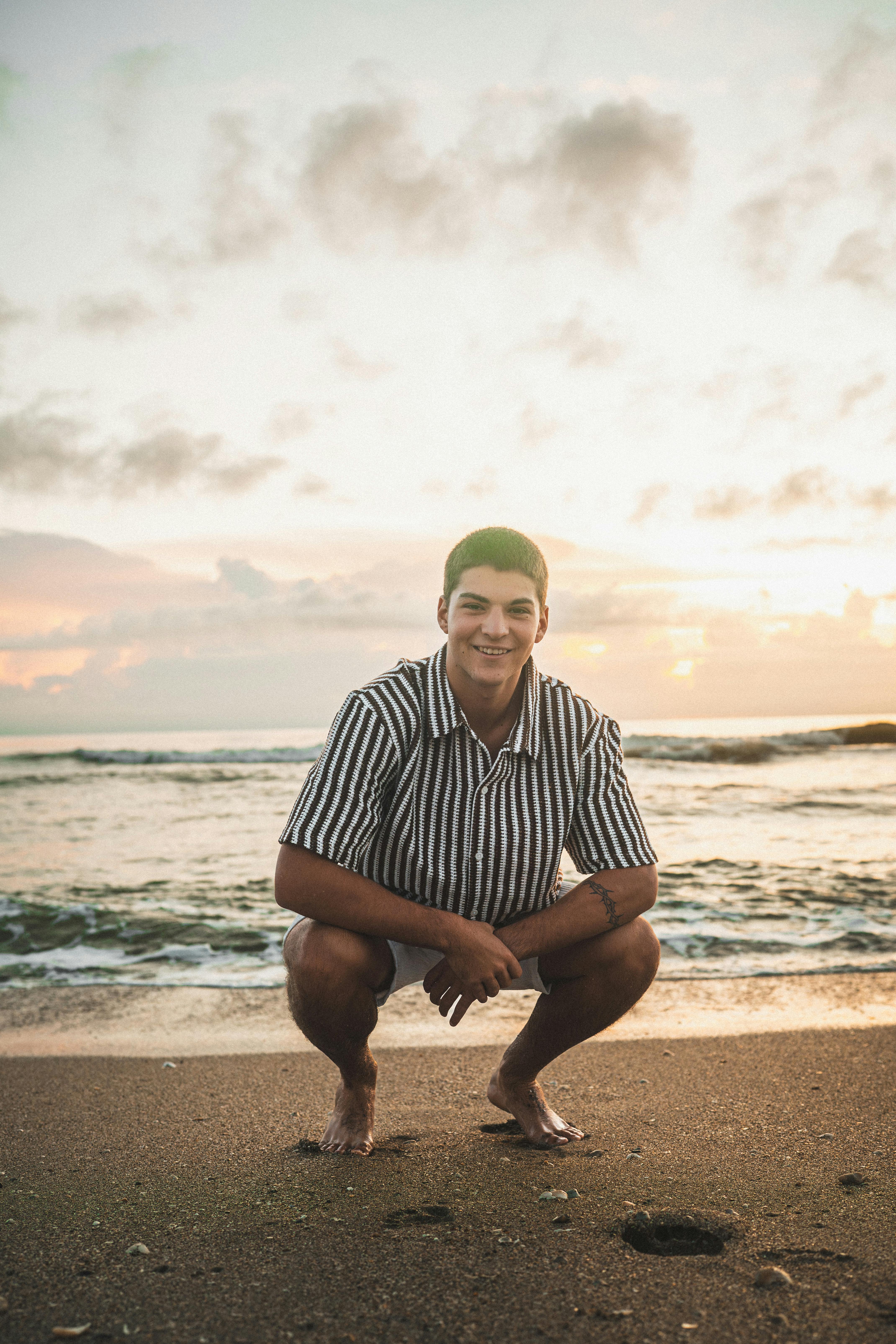Gratis Un joven sonriente con camisa de rayas posa en la playa de Melbourne durante una impresionante puesta de sol. Foto de stock