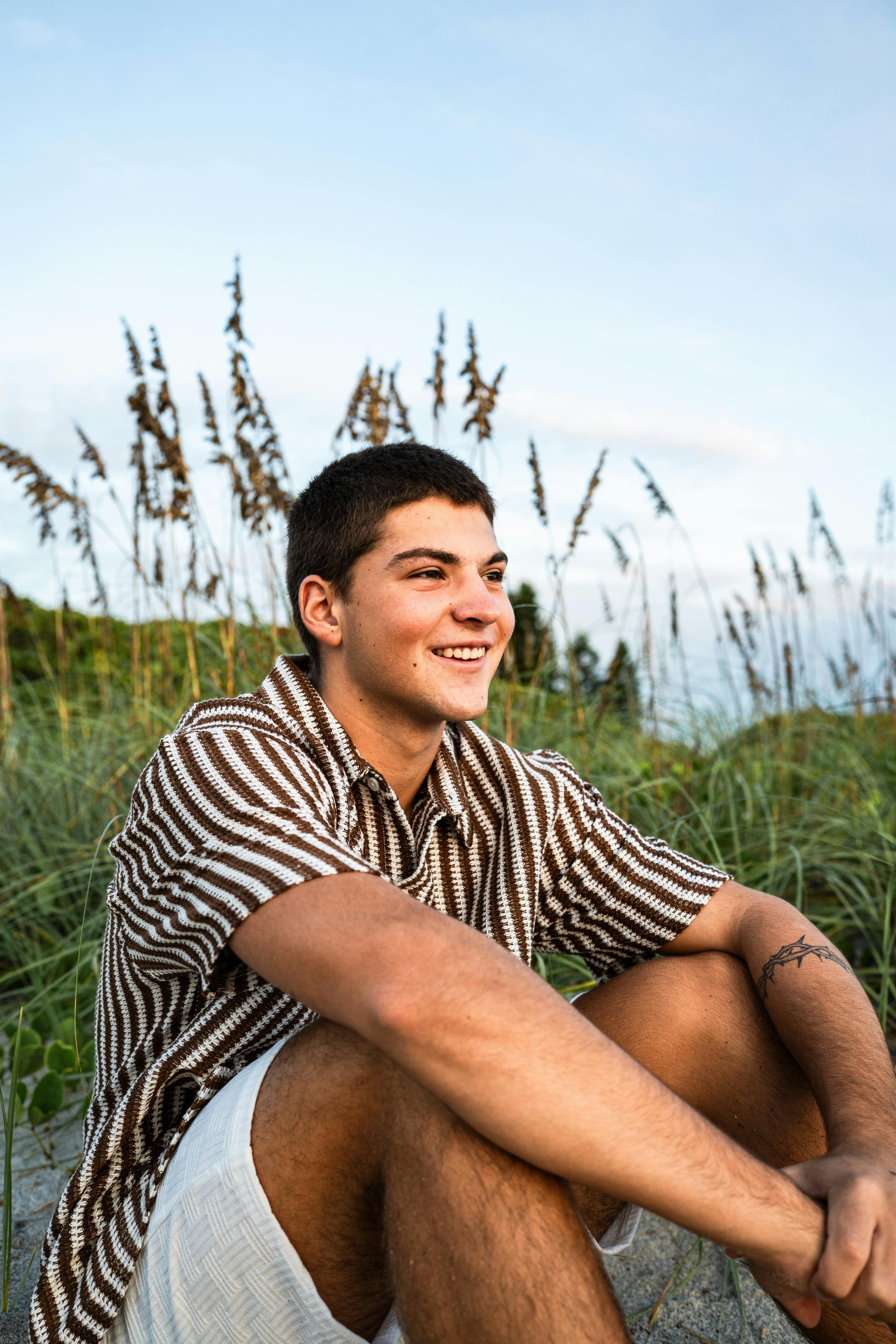 gratis Een lachende jongeman met kort haar zit op de duinen van Melbourne Beach, omringd door zeegras. Stockfoto