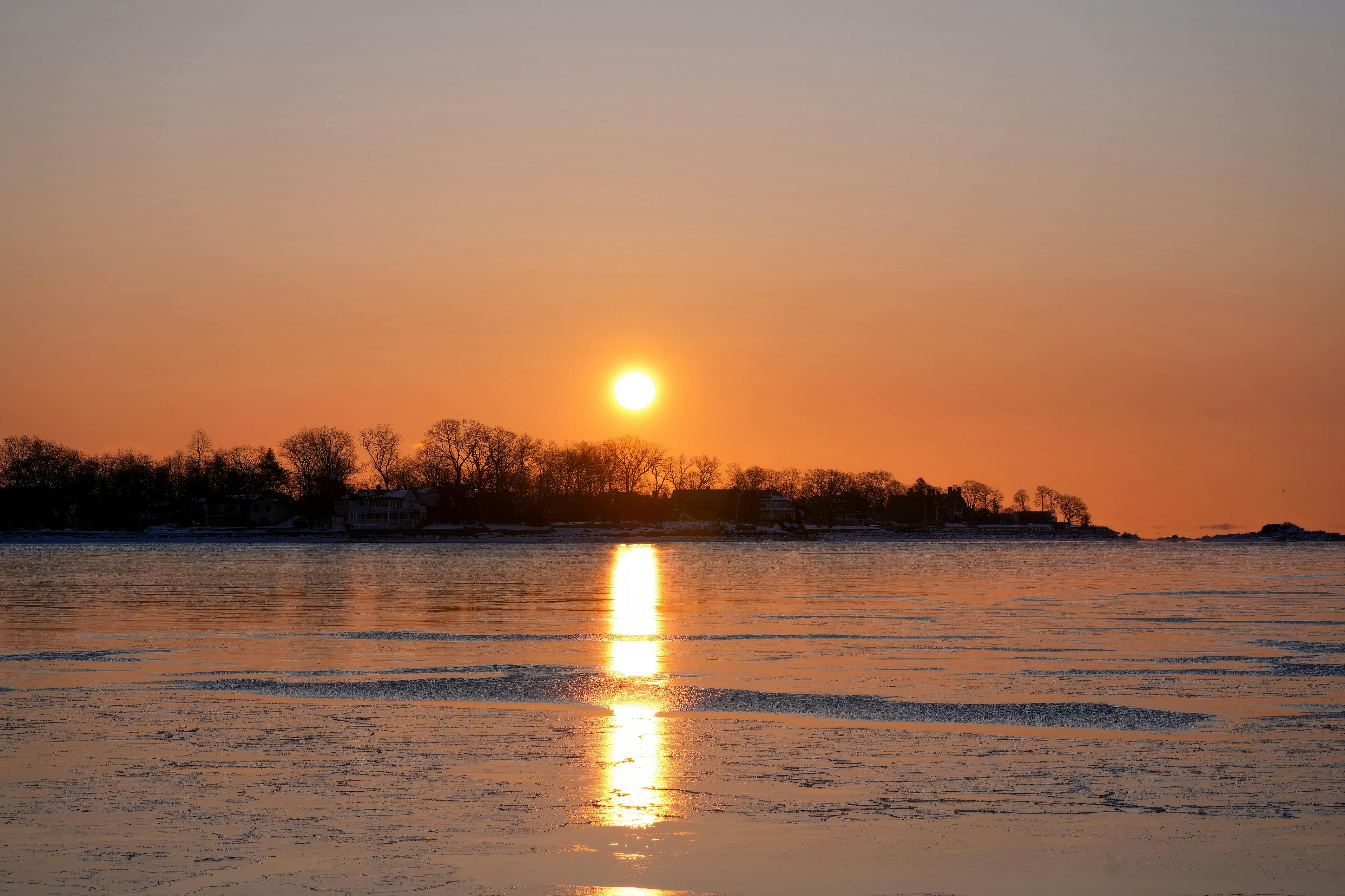 бесплатная Спокойный зимний закат, отражающийся в замерзшем озере, на фоне силуэтов деревьев. Стоковое фото