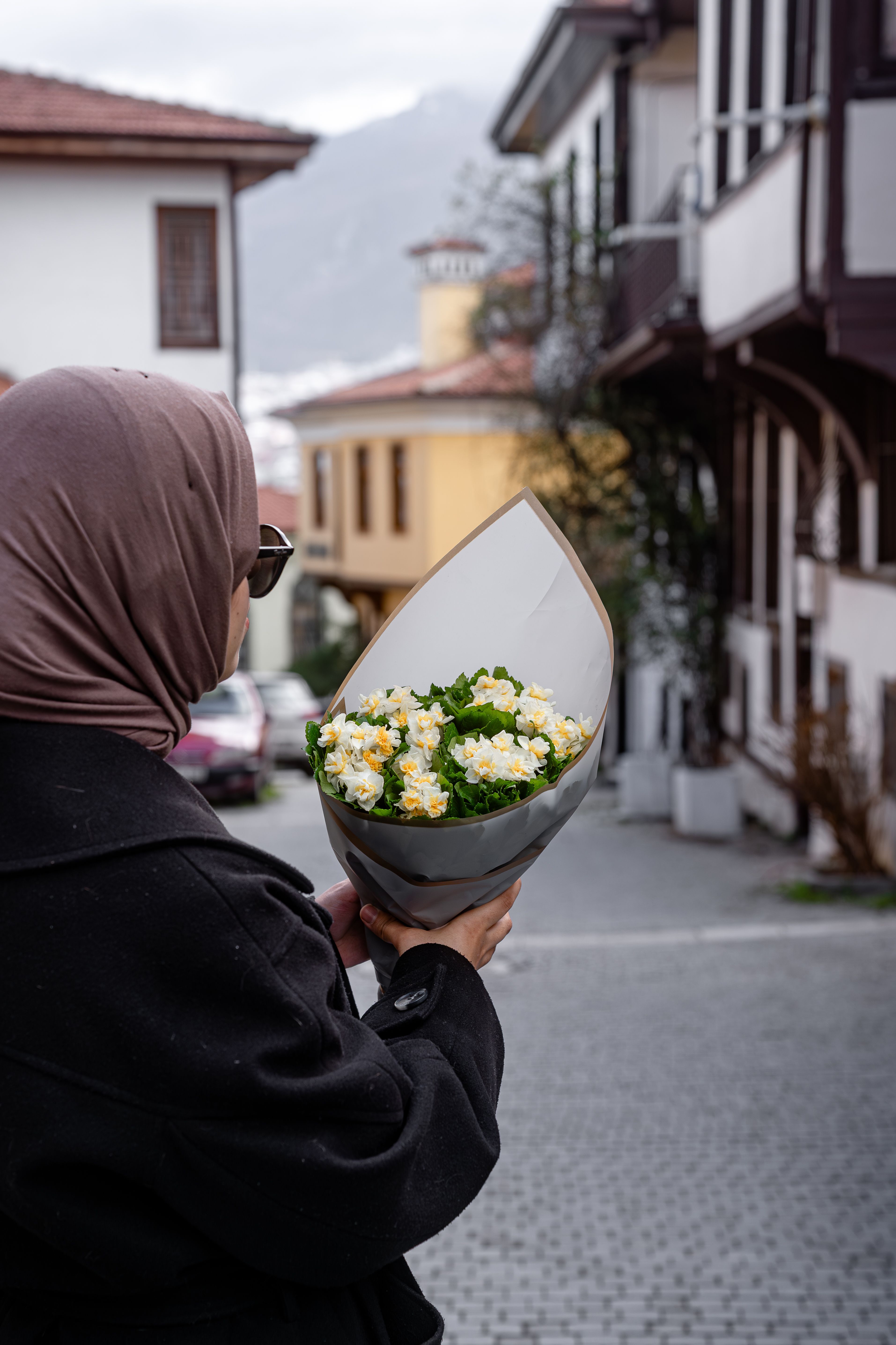 A woman in a hijab holds a bouquet of flowers on a peaceful street view with classic architecture.