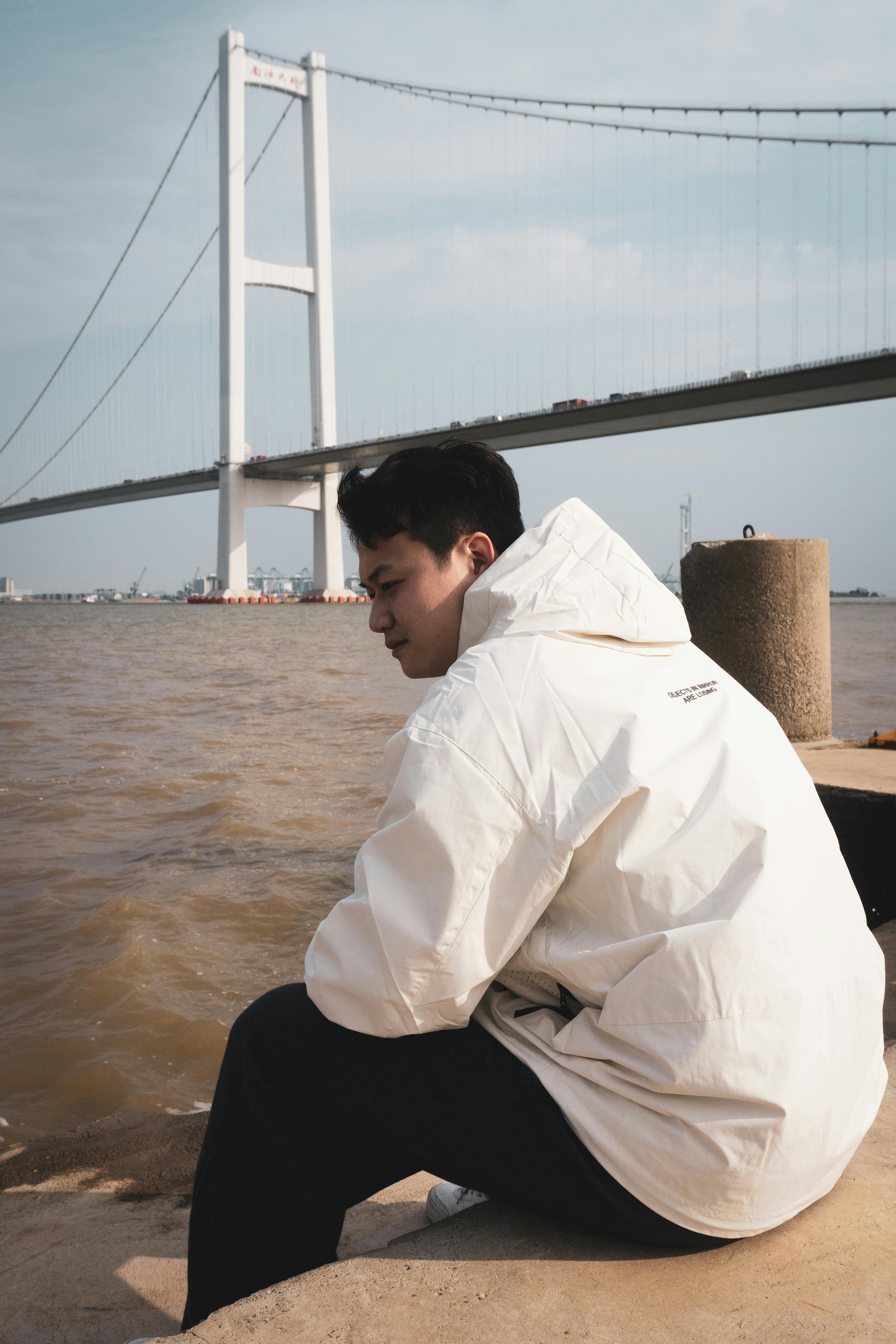 ingyenes Egy fehér zakós fiatalember ül egy folyó partján, egy nagy függőhíd közelében, Kantonban, Kínában. Stockfotó