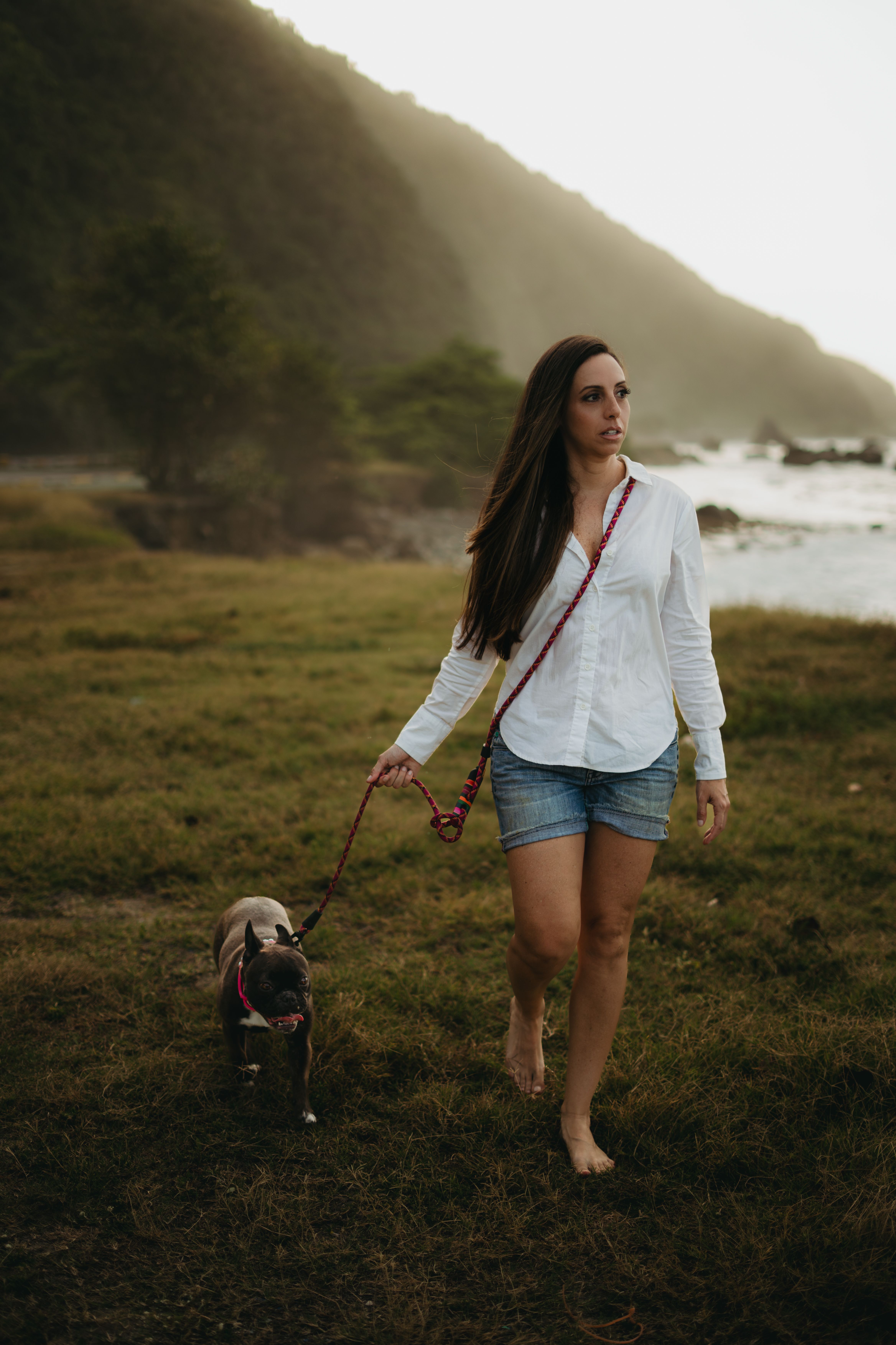 A woman walking her dog on a grassy coastal path at dusk, with hills and the ocean in the background.