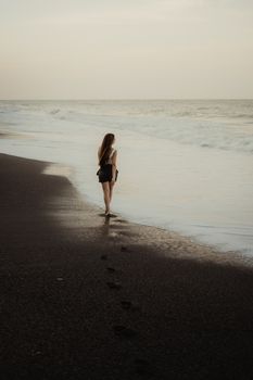 A pensive young woman walks along an empty beach at dusk, leaving footprints in the sand.