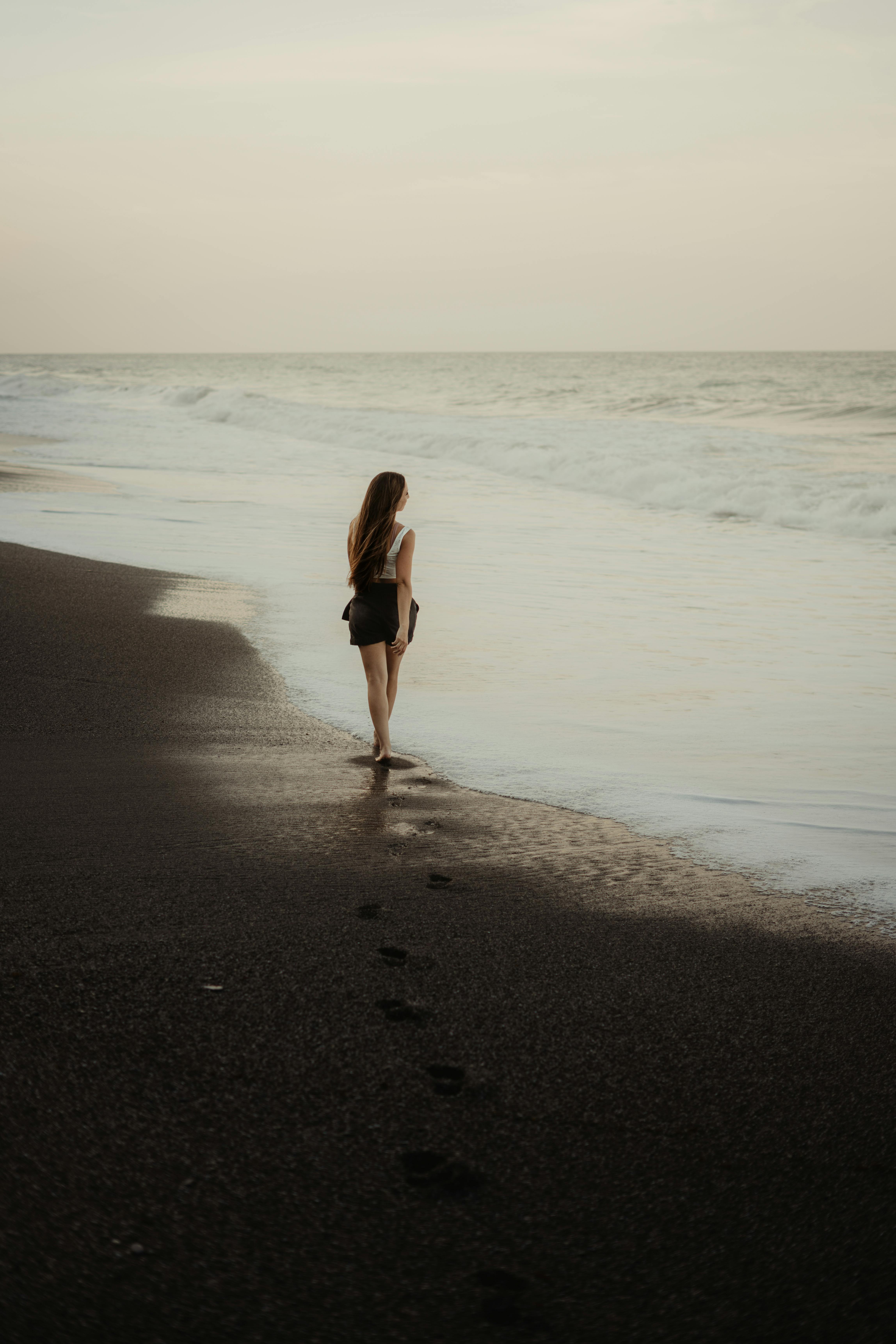 Miễn phí Một cô gái trẻ trầm tư bước dọc theo bãi biển vắng vẻ lúc hoàng hôn, để lại những dấu chân trên cát. Ảnh lưu trữ