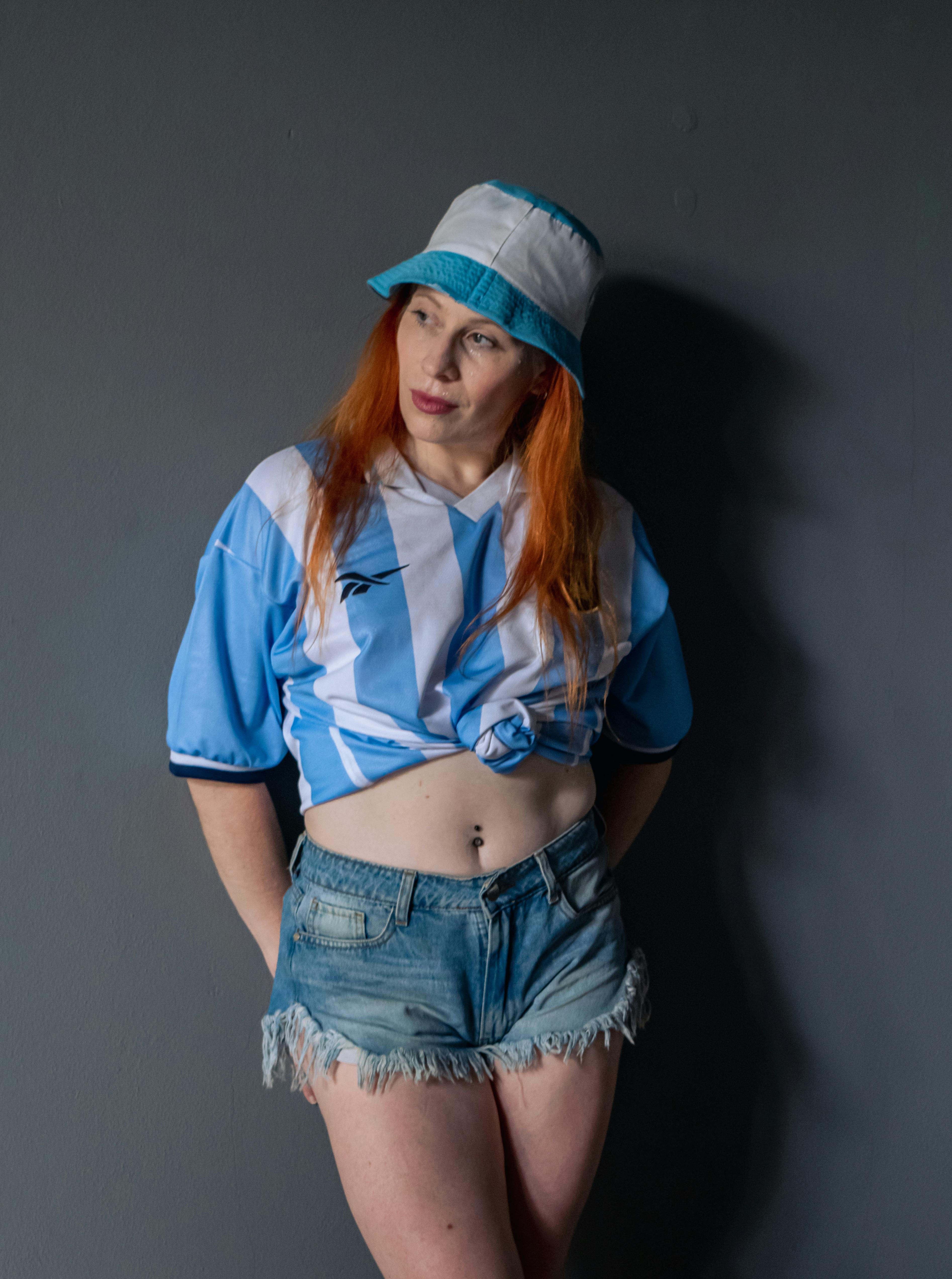 ingyenes Kék-fehér sportmezben és farmernadrágban lévő nő pózol beltérben, szürke háttér előtt. Stockfotó