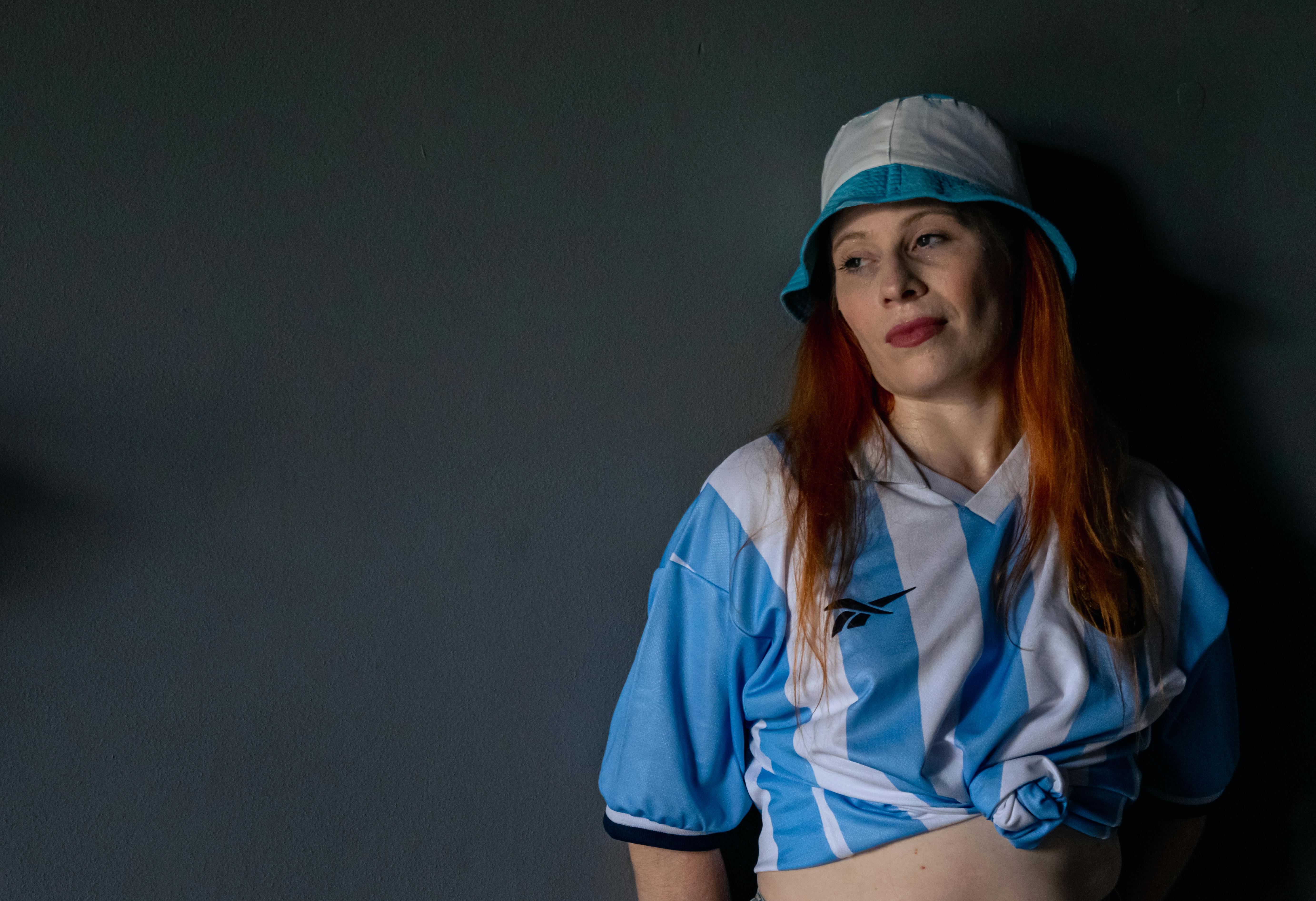 Zadarmo Žena v argentínskom futbalovom drese a čiapke, opretá o stenu v interiéri. Fotka z fotobanky